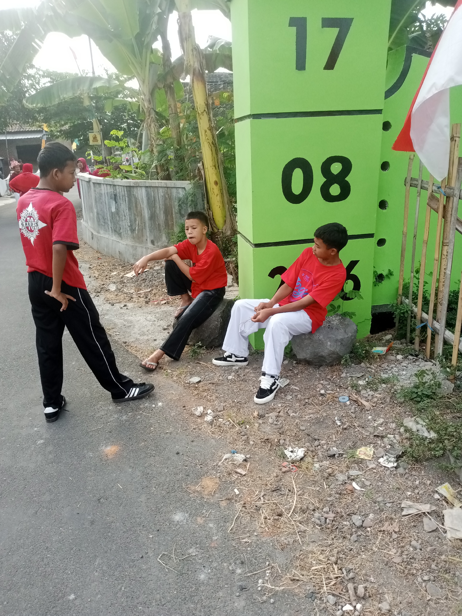 In Karangpakel, Indonesia, three young boys are present near a lime-green post with large black numbers "17" and "08" on it. One boy, wearing a red t-shirt with a white emblem and black pants, stands with his left hand on his hip, facing left. To his right, another boy in a red t-shirt and black pants sits on a large rock, looking to his left and gesturing with his right hand. A third boy, also in a red t-shirt and white pants, sits on a rock to the right, looking to his left. An Indonesian flag is visible near the green post. The ground is covered in gravel and dry grass. Banana trees and other vegetation are in the background, along with a concrete wall and a fence made of bamboo poles. The scene appears to be on a street or roadside.