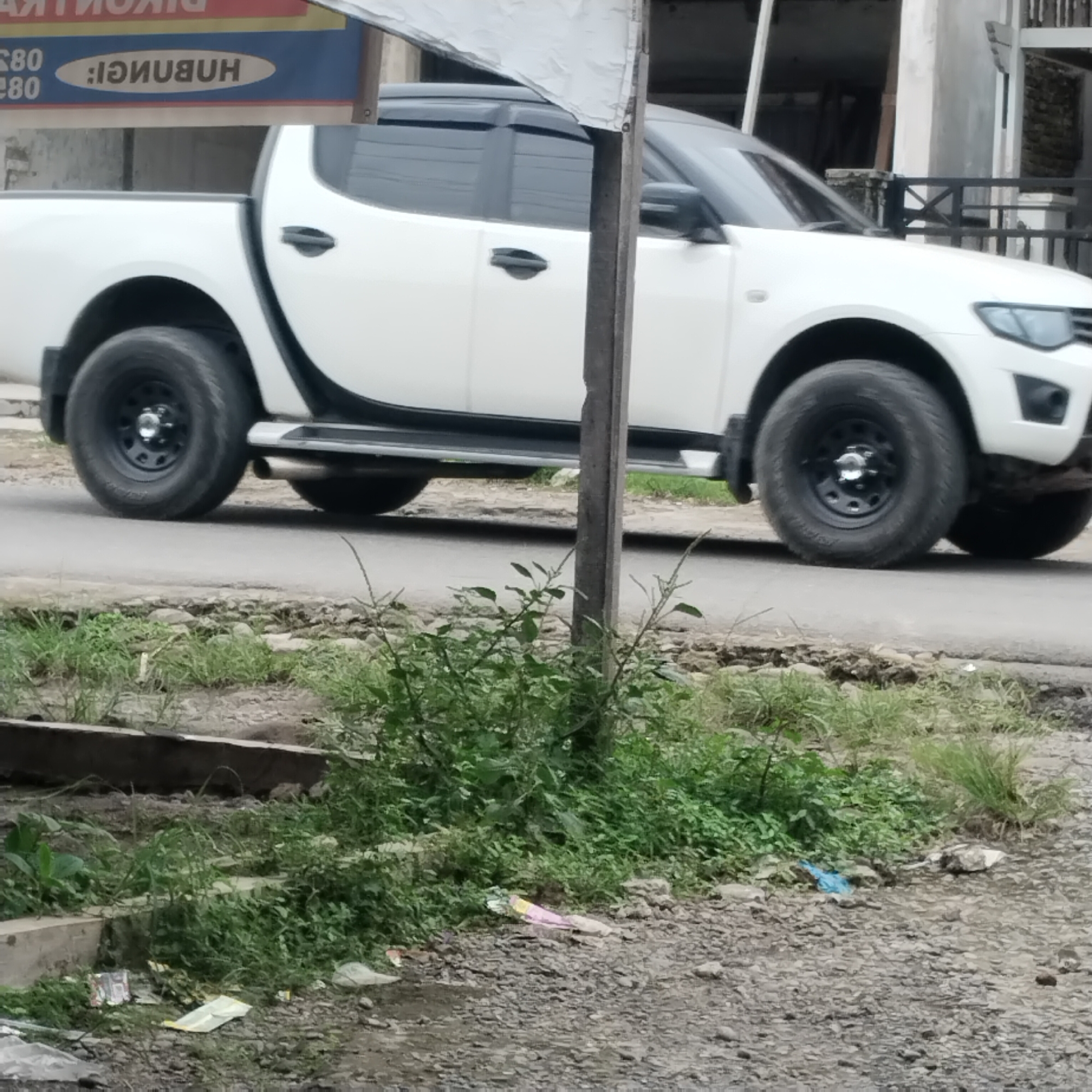 A white pickup truck with black trim is parked on a paved road. The truck's doors, fenders, and wheels are visible. The truck is parked in front of a building with a black metal fence. The area around the truck is overgrown with grass and weeds. There are several pieces of trash on the ground, including plastic bags and wrappers. The image appears to have been taken in Talun Kenas, Indonesia.