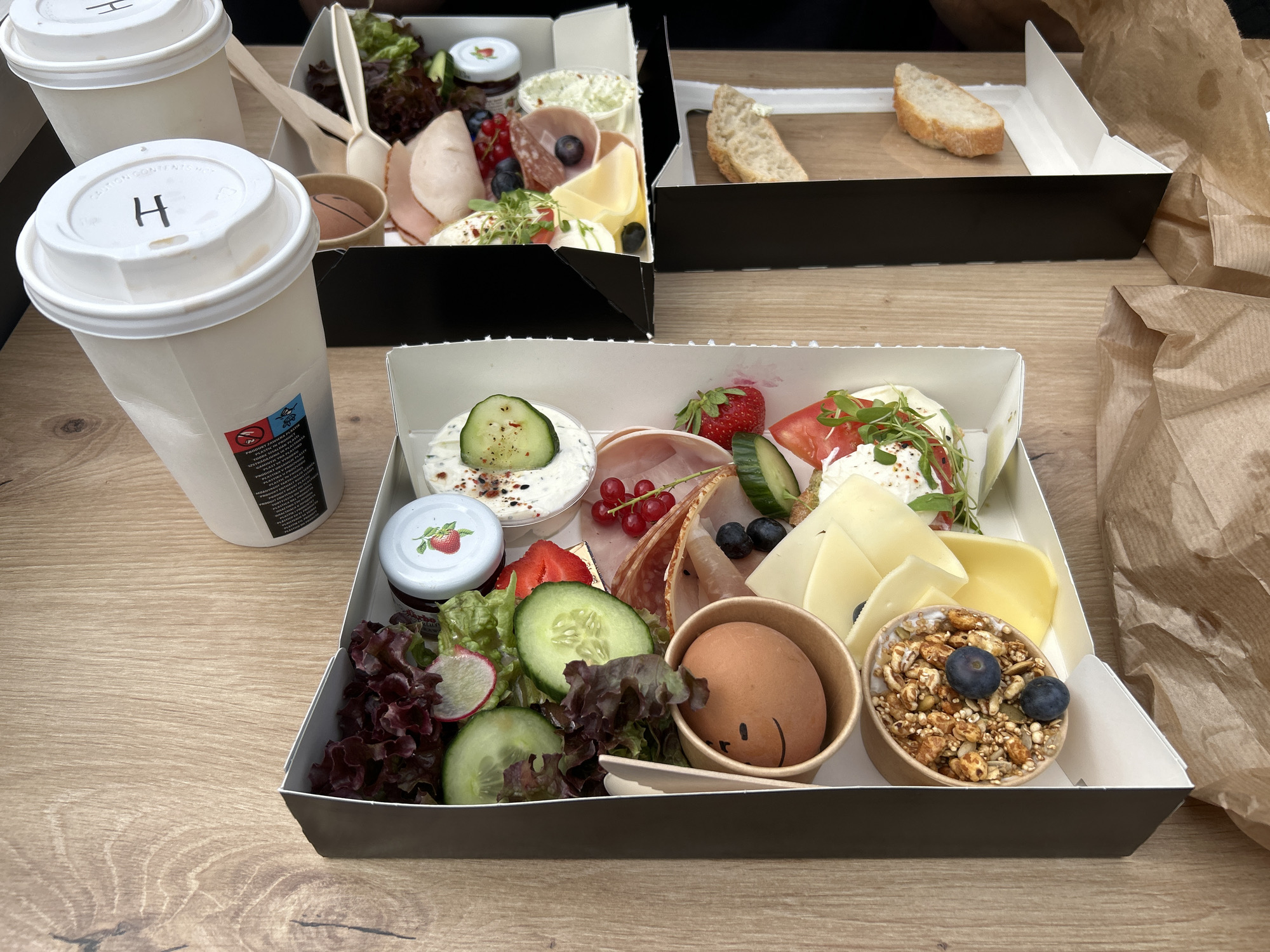 In an overhead shot of a wooden table, two takeout boxes of breakfast are presented. The box in the foreground is filled with various breakfast items, including a hard-boiled egg with a drawn smiley face, a small cup of granola topped with blueberries, slices of cheese, fresh berries, cucumber slices, lettuce, radishes, and what appears to be some type of cured meat. Next to this box, to the left, are two white paper cups with lids, one with the letter "H" drawn on it. The second takeout box, positioned behind the first and slightly to the left, contains a piece of bread and a small portion of salad with cured meat. Brown paper bags are visible on the right side of the frame. The lighting is natural and bright, indicating an outdoor or well-lit indoor setting.