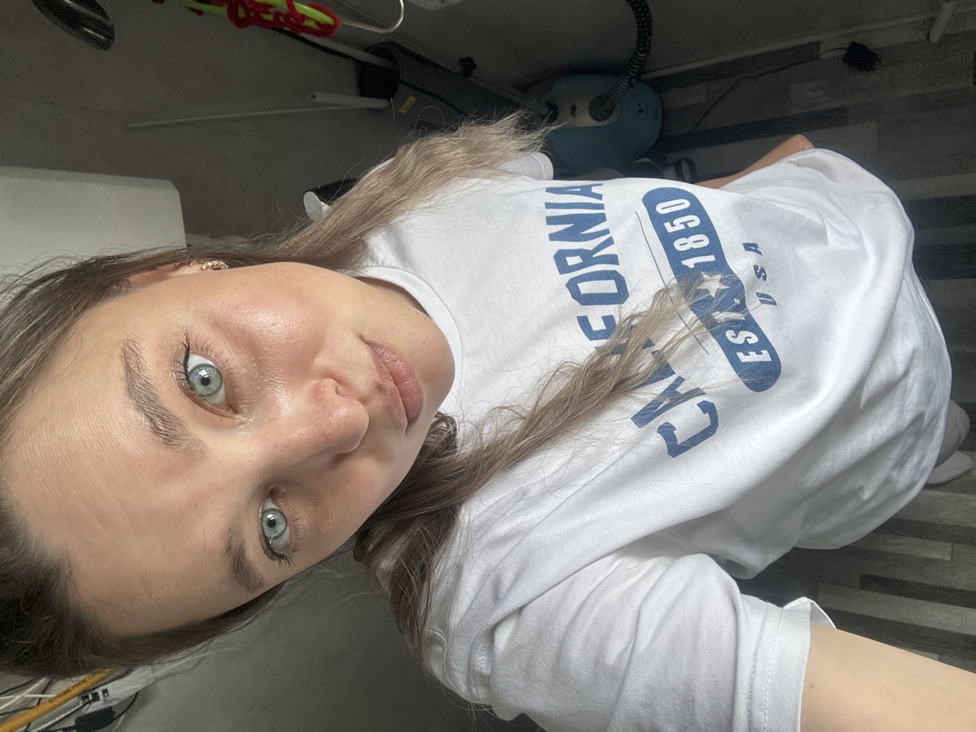 A young woman with blue eyes and blonde hair is taking a selfie in what appears to be a bathroom or utility room. She is wearing a white t-shirt with blue lettering that says "CALIFORNIA EST. 1850 USA". The background is somewhat cluttered with household items, including what looks like a vacuum cleaner. The lighting is bright and natural. The floor is tiled with a wood-grain pattern.
