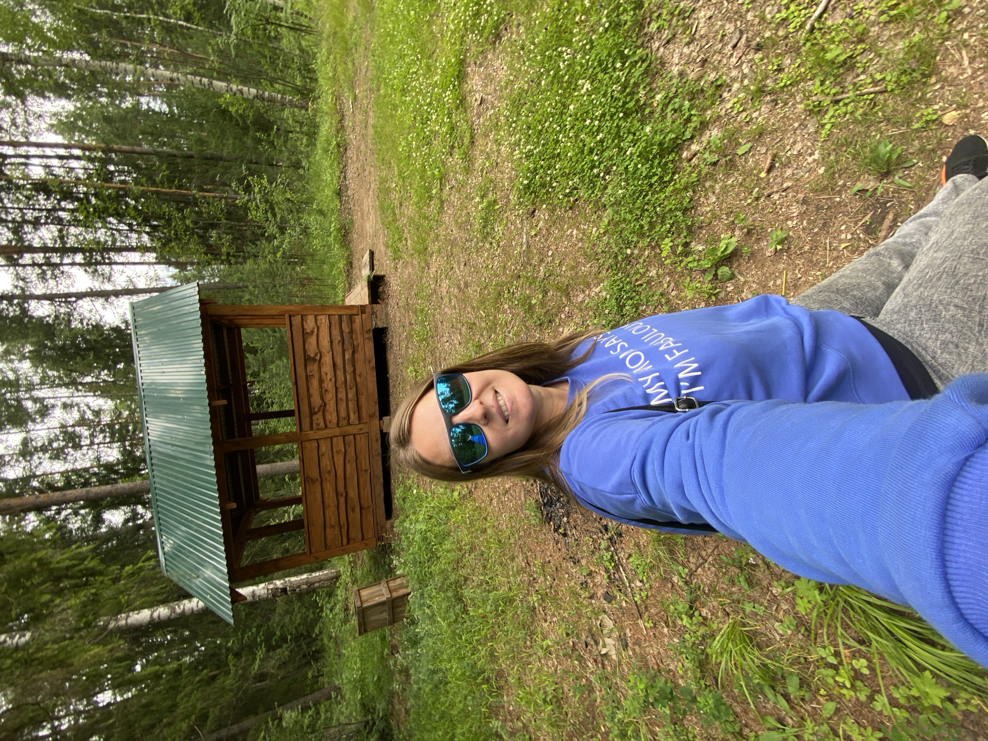 A woman is taking a selfie outdoors in a forest near Noviy, Russia. She is smiling and wearing blue sunglasses, a blue sweatshirt with text on it, and grey pants. Behind her, there is a wooden gazebo with a green metal roof and trees. The ground is covered in grass and dirt.