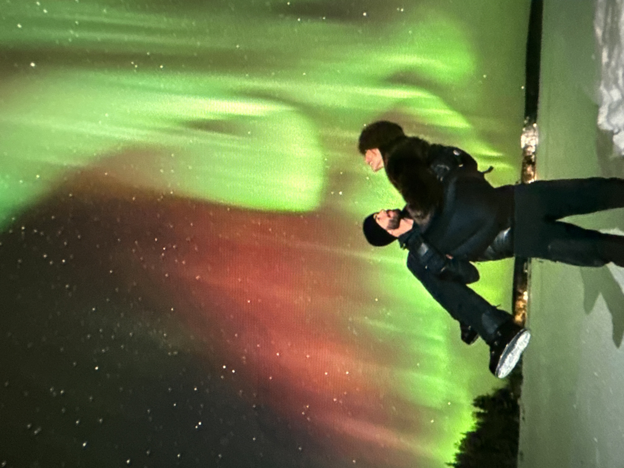 A couple, dressed in dark winter clothing, is posed against a backdrop of the aurora borealis. The aurora displays bands of green and red with numerous stars in the black sky. The location appears to be near trees and possibly a body of water or snowy field. Location: Unknown, Unknown.
