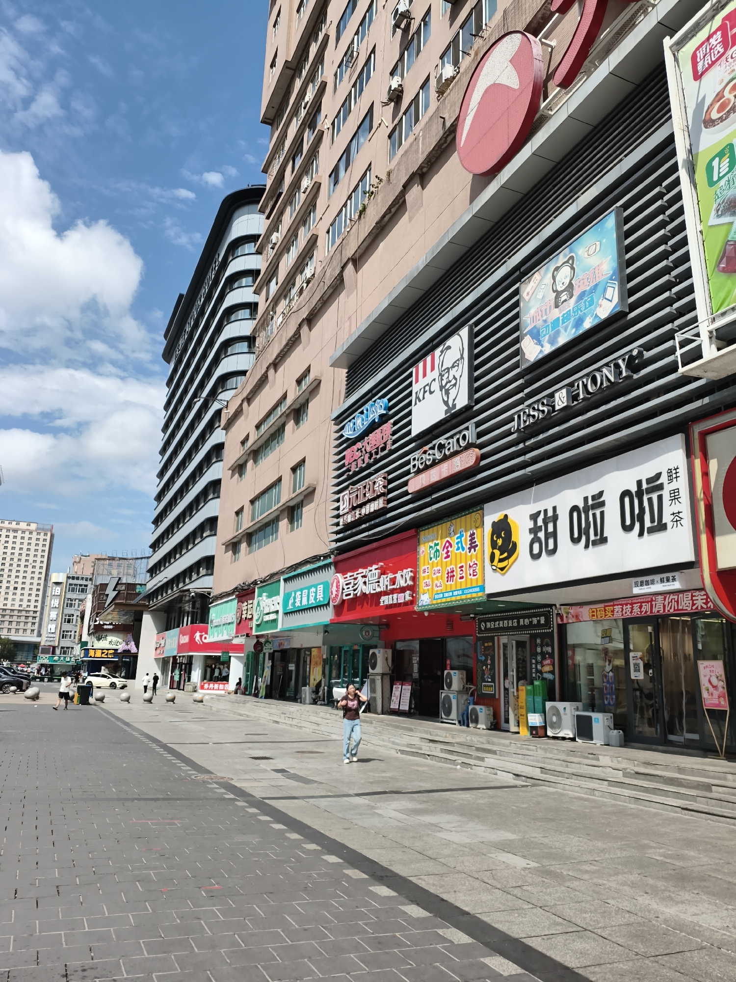 The image captures a street scene in Mudanjiang, China, on a clear day with scattered clouds. A multi-story building dominates the right side of the frame, displaying various storefronts and signage. Among the visible signs are "KFC", "JESS & TONY", and "BesCarol". Below these, a large white sign features black Chinese characters that appear to be advertising a beverage or food item, possibly "sweet tea". To the left of the main building, another tall, modern building with a curved facade is visible, along with other commercial structures further down the street. The foreground shows a paved walkway with a distinct pattern of dark gray rectangular stones. Several people are present: a woman walks on the right side of the walkway, talking on her phone; a person in a white shirt and blue pants stands in the middle of the scene, looking at their phone; and other individuals are seen walking or standing near the shops in the background. Cars are parked along the curb on the far left of the image. The environment appears to be a commercial district with a mix of architectural styles.