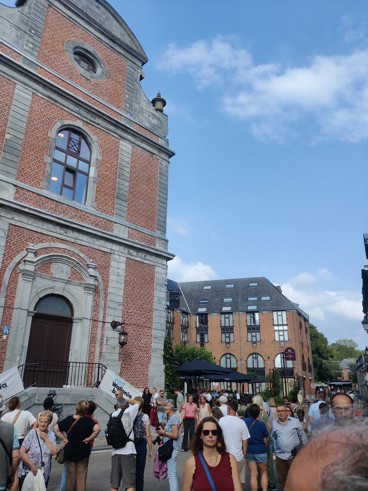 The image depicts a street scene in the city of Marche-en-Famenne, Belgium, likely during a public event. In the foreground, a crowd of people are gathered, some looking towards a building on the left, others in conversation or walking. The building on the left is a large brick structure with stone accents and arched windows. Two banners are visible on the steps of this building; one reads "VISIT Wallonia b." and the other is partially obscured.
Further down the street, on the right side of the image, is a multi-story brick building with many windows, appearing to be an apartment building or hotel. A sign on this building reads "HOTEL RESTAURANT" and below it, "Le Comte de Meeus". Outdoor seating areas with black umbrellas suggest restaurants or cafes. The sky is blue with scattered white clouds. The overall impression is a bustling urban environment on a pleasant day.