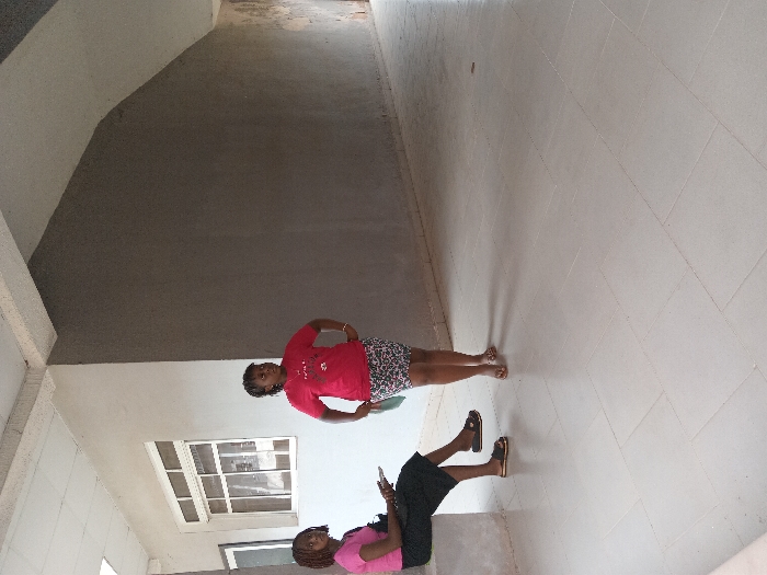 This image depicts two adult females within an indoor or semi-enclosed common area of a building in Ogboko, Nigeria. The floor is laid with light-colored square ceramic tiles. Walls are painted in two distinct tones: a lighter grey/off-white on the left, which includes white-framed windows, and a darker grey on the right. Overhead, a suspended ceiling with white tiles and structural beams is visible, alongside angled architectural features.
On the left, one female is seated on a light-colored concrete bench. She is dressed in a pink short-sleeved top, a dark skirt, and light brown flat sandals. She holds a dark electronic device in her hands and directs her gaze downwards. A dark bag rests on the bench beside her.
Standing slightly forward and to the right is a second female. She wears a red short-sleeved t-shirt with a graphic design, a patterned short skirt, and light brown flat sandals. Her hands are positioned on her hips, and she holds a light-colored object in her left hand, looking towards the left side of the frame. There is no direct interaction observed between the two individuals.