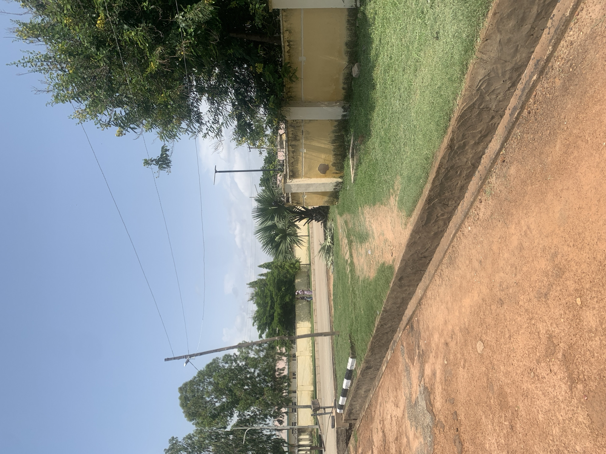 This image depicts an outdoor scene, likely in Jalingo, Nigeria. The foreground is dominated by a rough, reddish-brown dirt path bordering a drainage channel. To the left of the channel is a strip of vibrant green grass.
Beyond the grass, a light yellow wall runs along the property line, with some signs of wear and tear. Trees with lush green foliage are visible behind the wall, some reaching over it. In the middle ground, a paved road is visible, with a person on a bicycle or motorcycle riding away from the camera. Further down the road, more trees create a dense green backdrop.
Power lines crisscross the clear blue sky, supported by utility poles. The lighting suggests it is daytime, likely mid-morning or afternoon, with bright sunshine casting shadows. The overall atmosphere is ordinary and depicts a common street scene in a tropical or subtropical climate. No visible text is present in the image.