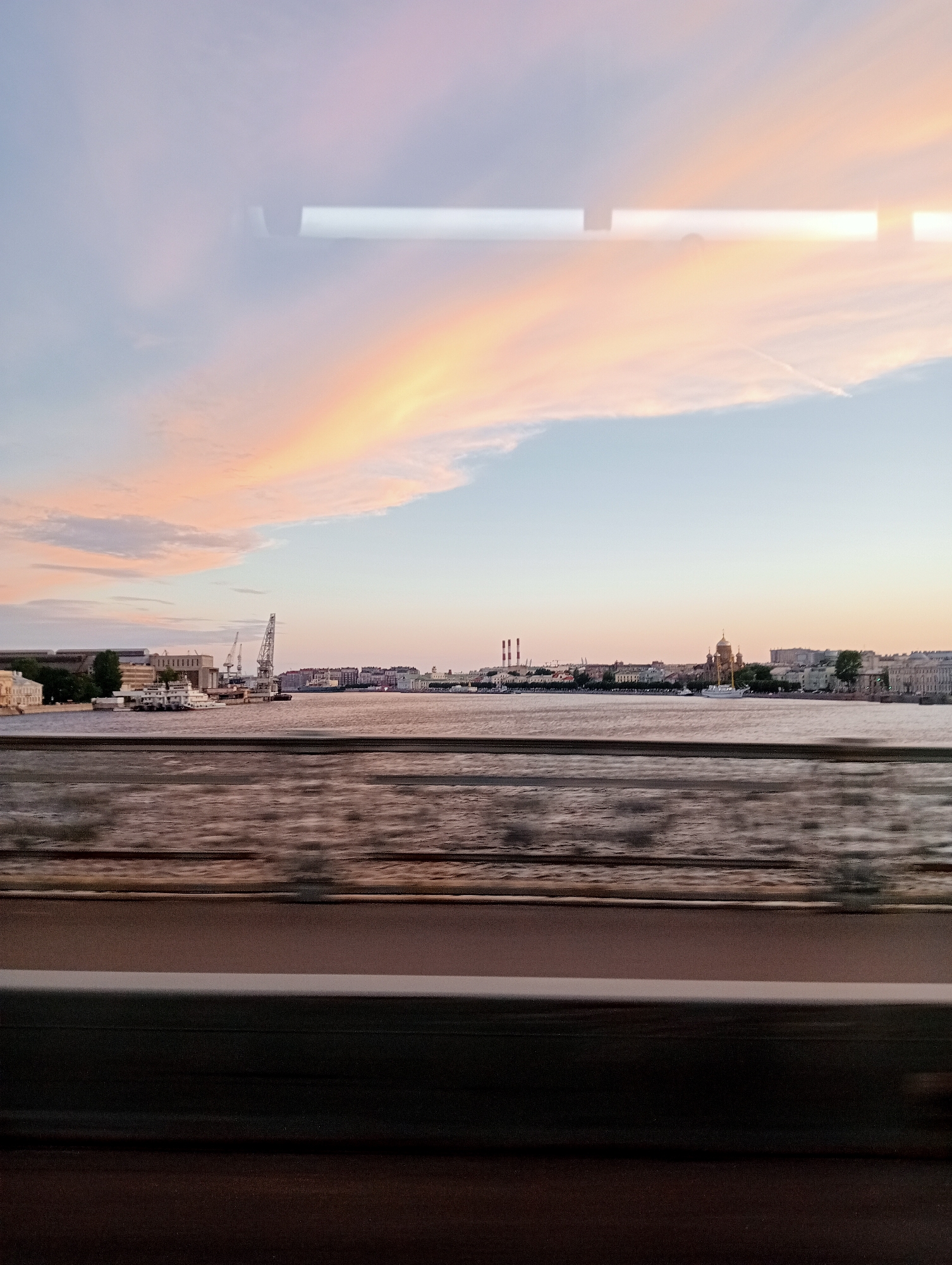 This is a photograph taken from a moving vehicle, likely a train or bus, looking out at the Neva River in Saint Petersburg, Russia, during sunset. The sky is a gradient of soft pinks, oranges, and blues, with wispy clouds. In the distance, across the river, the city skyline is visible. On the left side of the river, there are buildings, cranes, and what appears to be a ship or dock. Further along the skyline, there are more buildings, industrial chimneys, and a prominent domed building, likely St. Isaac's Cathedral, with a sailboat in front of it. The foreground shows the blurred interior of the vehicle, with a dark horizontal bar dominating the bottom portion of the image, suggesting the presence of a window frame or some other obstruction. The water of the Neva River is choppy and reflects the colors of the sky. The overall impression is one of a fleeting glimpse of the city's picturesque waterfront at dusk.