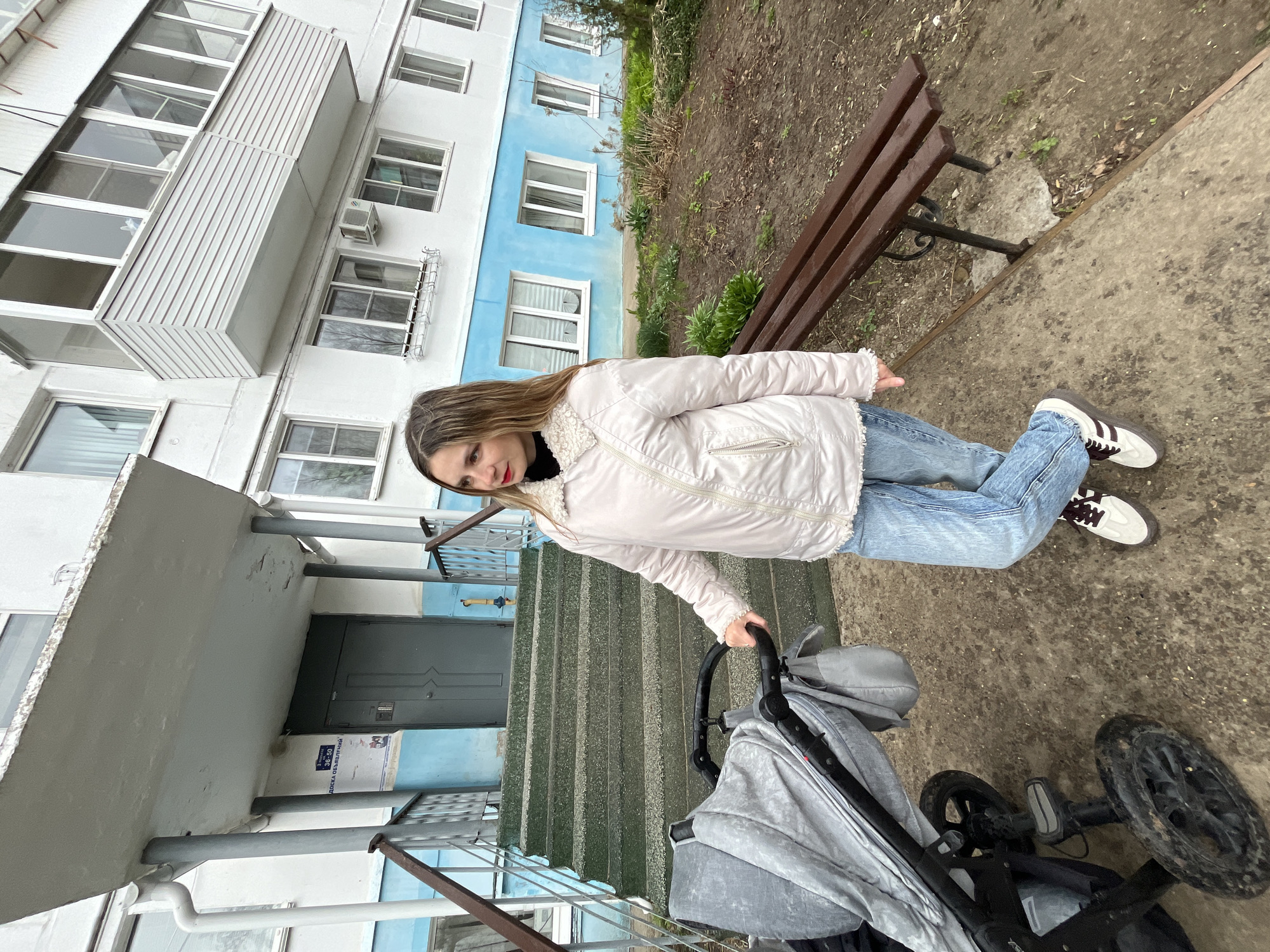 A young woman wearing a light-colored jacket and blue jeans is standing on a concrete walkway in front of an apartment building. She is holding the handle of a grey baby stroller. Behind her are stairs leading up to the entrance of the building. A wooden bench sits to the right of the walkway, next to a small patch of garden. The building has a white and blue facade with multiple windows. A forensic examination of the location using metadata from the image and Google Street View indicates it is located in the city of Chaikovsky, Russia.