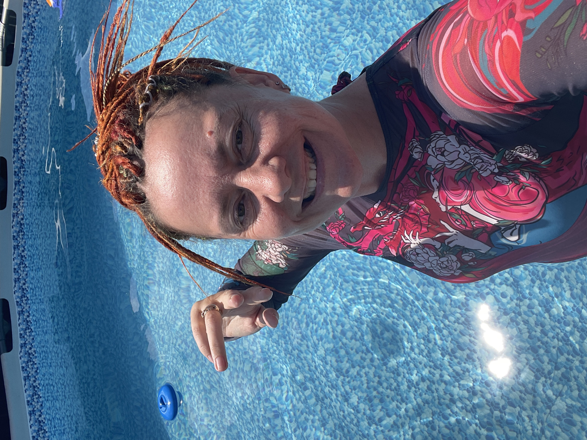 The image shows a woman with braided red hair standing in a swimming pool. She is wearing a long-sleeved rash guard with a floral and dragon-like print. Her left hand is raised, with her index finger pointing. The pool water is clear and blue, with sunlight reflecting on the surface. There is a blue pool float visible in the water on the left side of the image. The background shows the edge of the swimming pool with a blue and white tile pattern. There is no indication of the city of Orenburg, Russia, in the image.