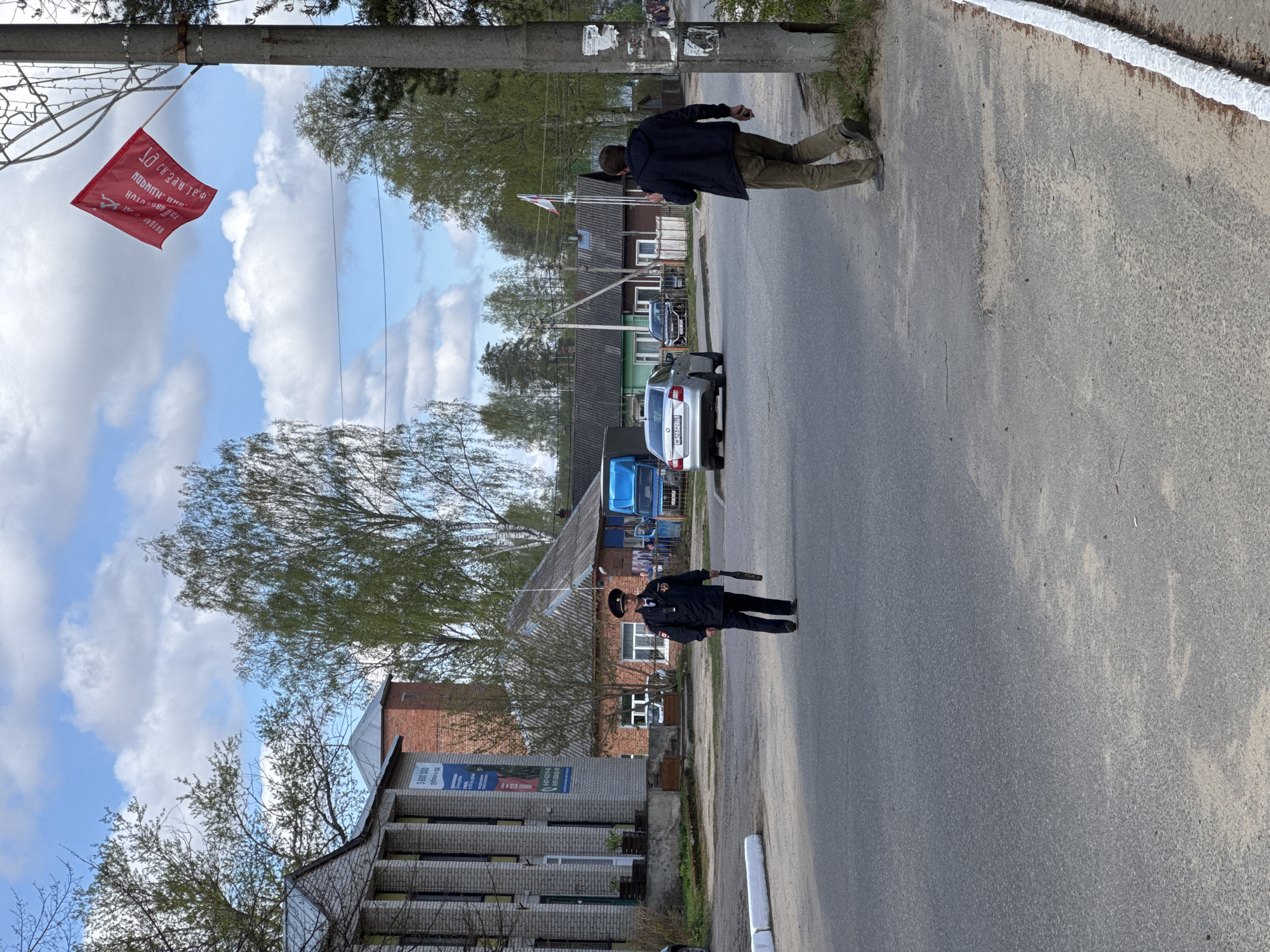 The image depicts a street scene in Chuprino, Russia. A man in civilian clothes is walking down the street, seemingly interacting with a police officer in uniform. The officer is holding what appears to be a metal detector. A silver car and a blue van are parked further down the street. In the background are residential buildings, and a red flag with white text hangs overhead. The sky is partly cloudy.