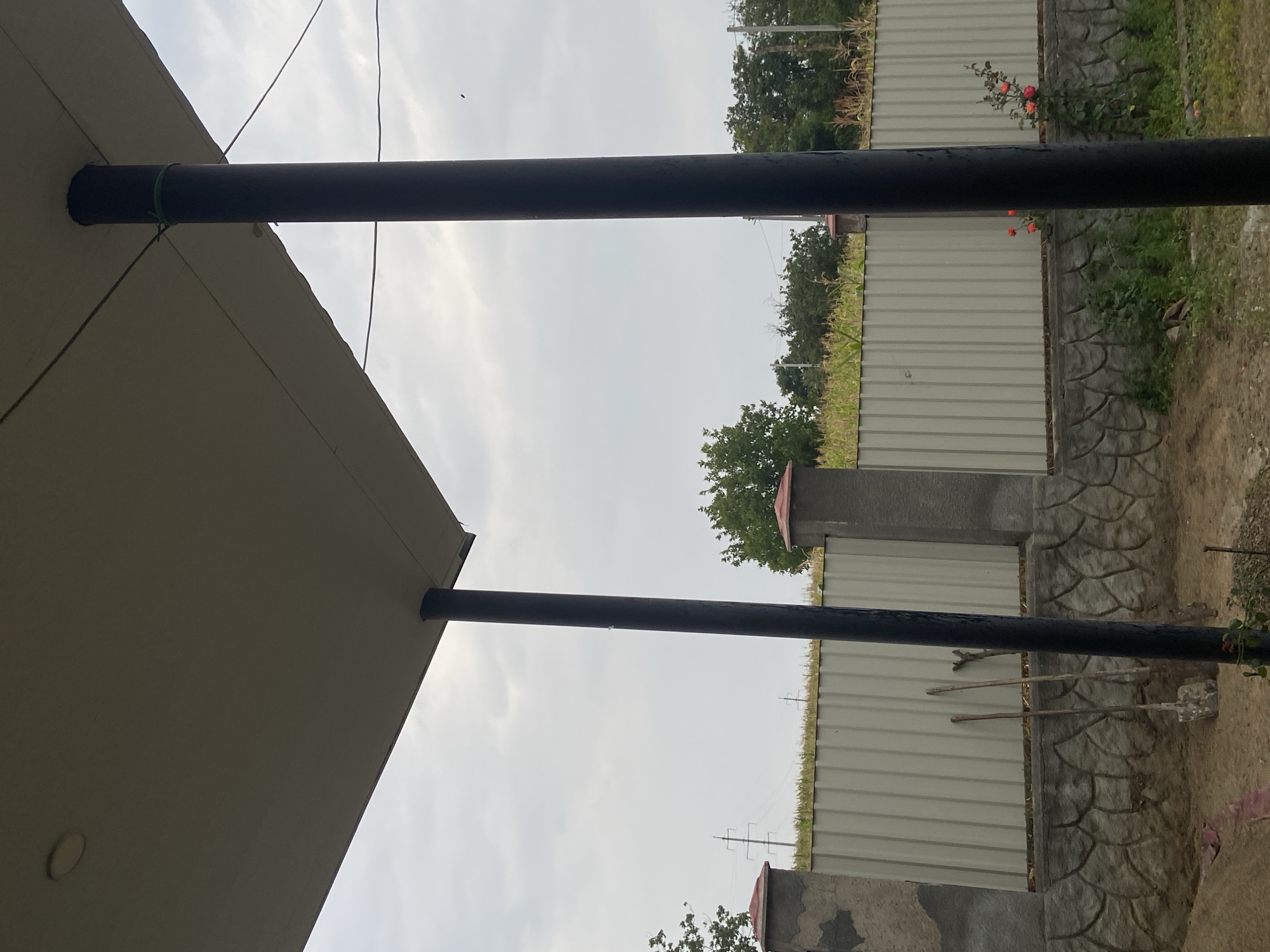 An outdoor, eye-level, medium shot captures a scene under a large, light-colored roof supported by two thick, dark poles. The sky above is overcast and pale. In the middle ground, a pale gray corrugated metal fence runs horizontally across the frame. The fence is built on top of a gray stone base that is textured to resemble interlocking stones. Two garden tools, a shovel and a rake, lean against the fence. The foreground shows a dirt area, possibly a construction site, with some scattered debris and a pink cloth visible. Beyond the fence, there is a dense line of green foliage and trees. In the distance, power lines and utility poles are visible against the sky. A small cluster of red roses is blooming to the right of the frame, near the base of the right-hand pole. The overall scene suggests a rural or suburban outdoor area, possibly undergoing renovation or construction.