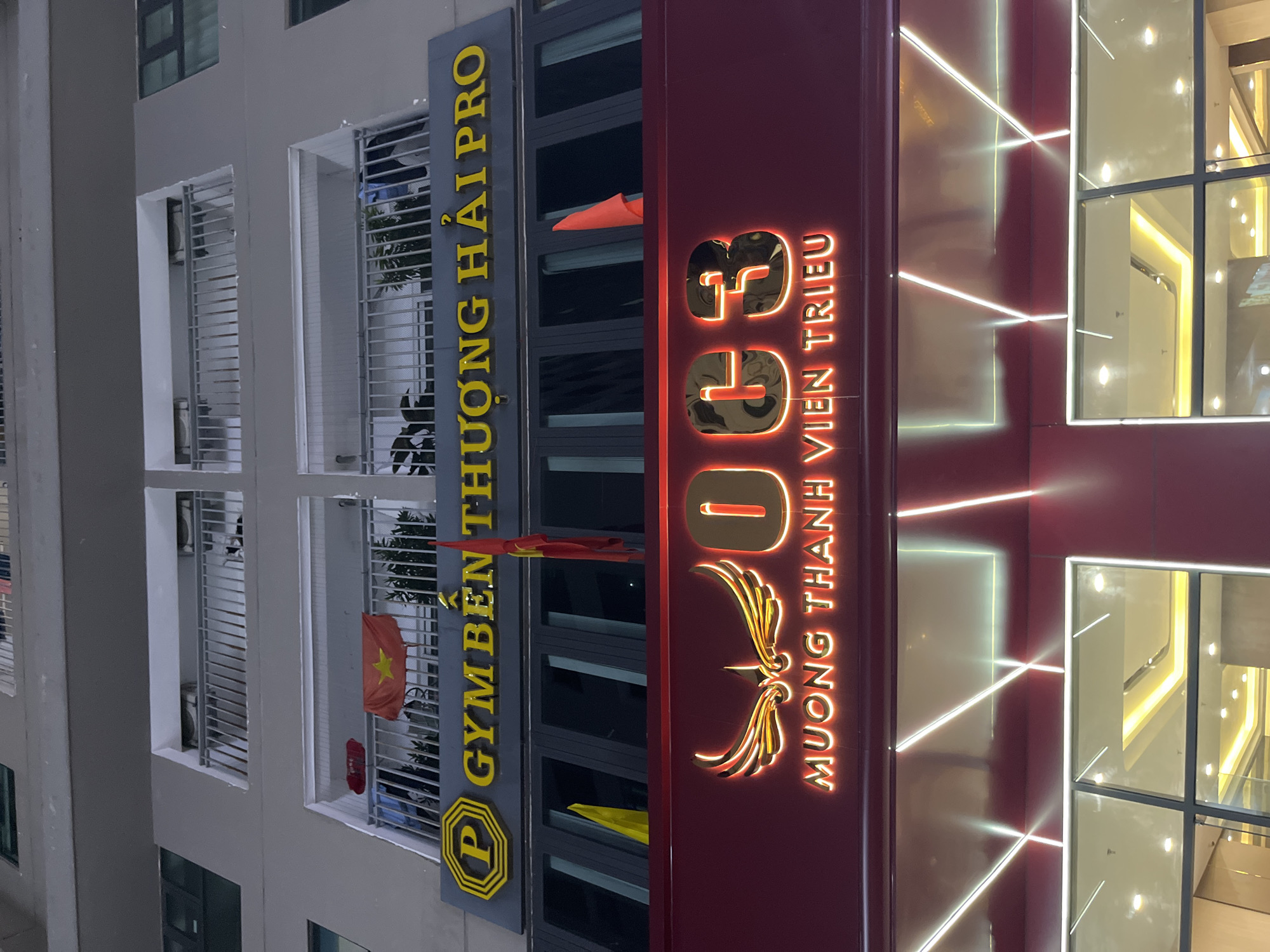 The image shows the facade of a building at night, likely a commercial establishment. A large yellow sign with the text "GYM BÊN THƯỜNG HẢI PRO" is prominently displayed. Below this, a red illuminated sign with stylized wings and the number "003" is visible, along with the text "MUONG THANH VIEN TRIEU". The building appears to be multi-story, with balconies on the upper floors. Two Vietnamese flags are visible on the balconies. The lower part of the image shows large glass windows reflecting internal lighting. The scene is set in the city of Bac Nha Trang Ward, Vietnam.