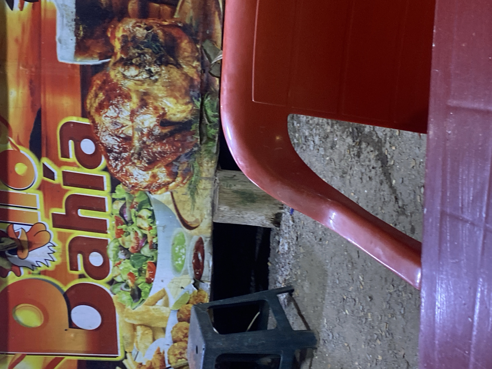 The image depicts a promotional poster for a restaurant, likely located in Juan Griego, Venezuela. The poster prominently features a roasted chicken dish, accompanied by salad, sauces, and fried food items. The restaurant name "bahía" is displayed vertically on the poster. A red plastic chair partially obscures the view, and a black plastic crate is visible near the bottom of the frame. The ground is a mix of dirt and gravel. No people are visible in the image.