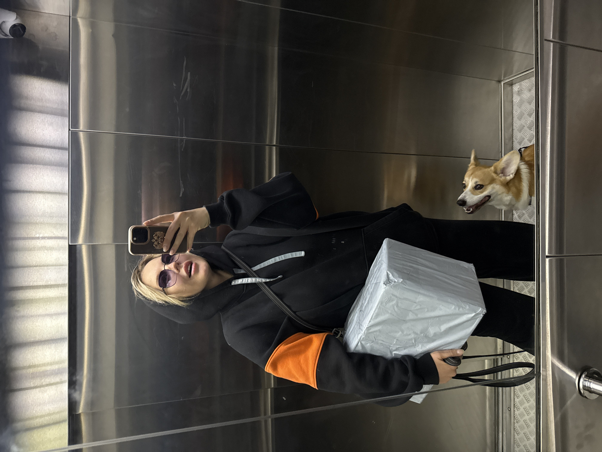 A woman takes a selfie in an elevator. She wears sunglasses and a black hoodie with orange sleeves. She holds a large, square, silver package with both hands. A corgi dog looks into the elevator from the outside. The elevator has stainless steel walls. There's a camera in the upper left corner. This image was taken in Krasnoyarsk, Russia.