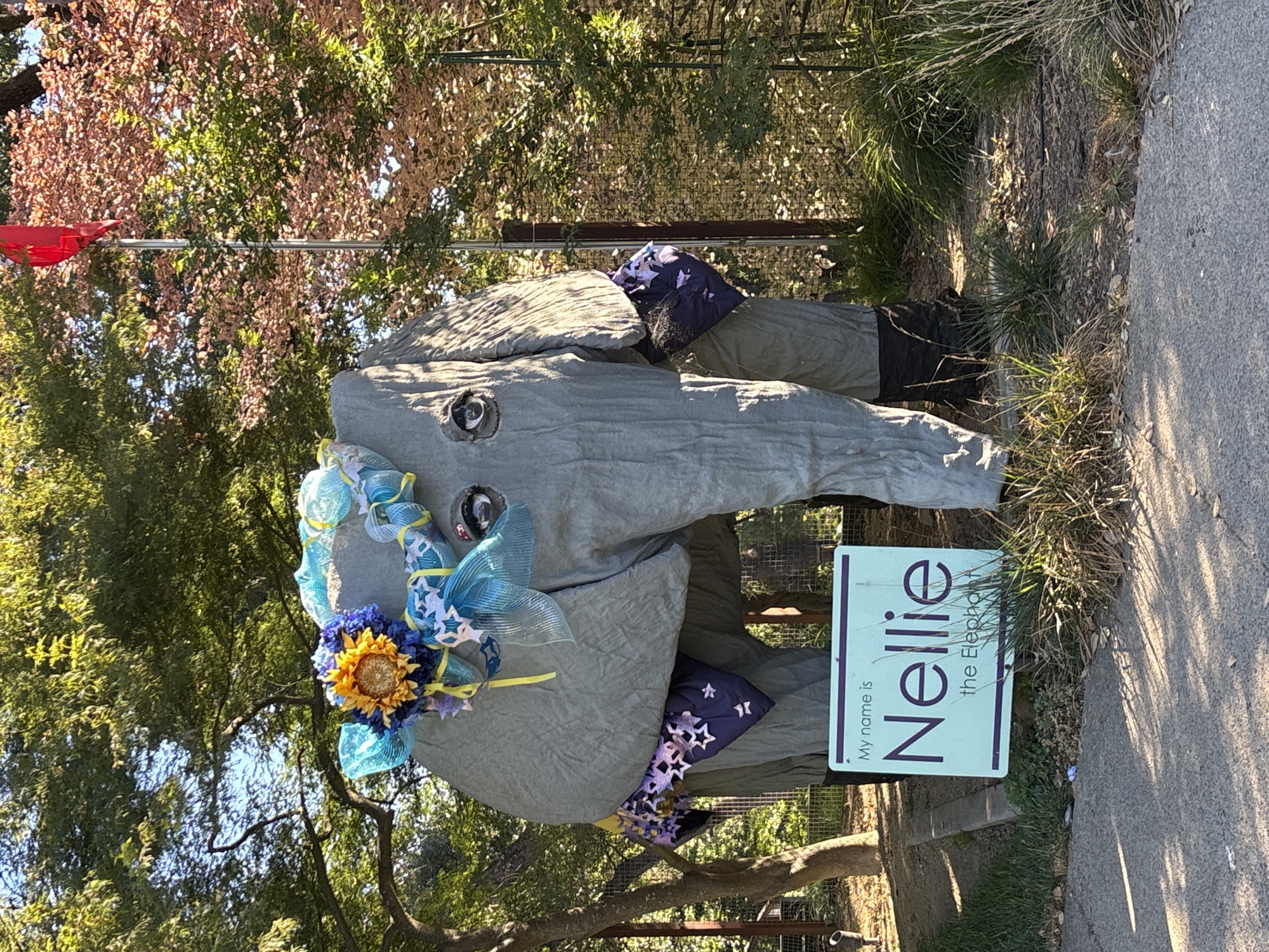A large, grey, elephant-shaped sculpture, identified by a sign as "Nellie the Elephant," stands outdoors in Fair Oaks, California. The sculpture is adorned with a colorful floral arrangement and blue mesh ribbons around its head, and a purple patterned cloth draped over its legs. A light blue sign in front of the sculpture reads "My name is Nellie the Elephant." The sculpture is situated in a grassy area with trees and a fence in the background. The lighting suggests it is daytime, with sunlight casting shadows. There are no people visible in the image.