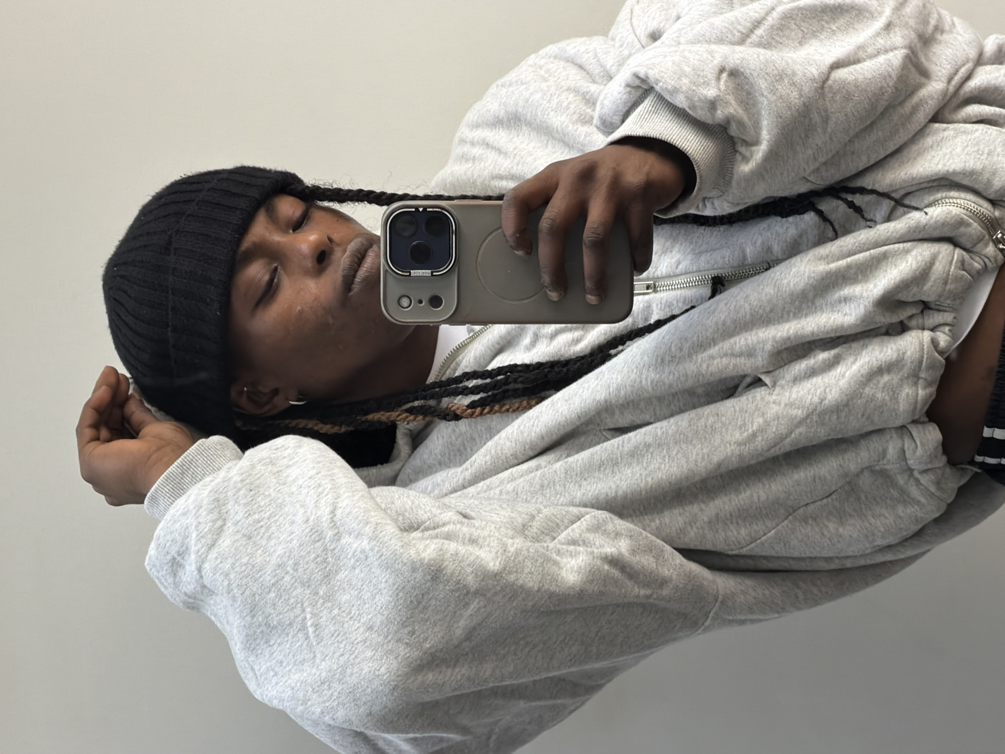 A person with braids is holding a smartphone in their hand, capturing a selfie. They are wearing a black beanie and a light grey, zip-up hooded sweatshirt. The background is a plain, light grey wall. The location is not discernible based on the image provided.