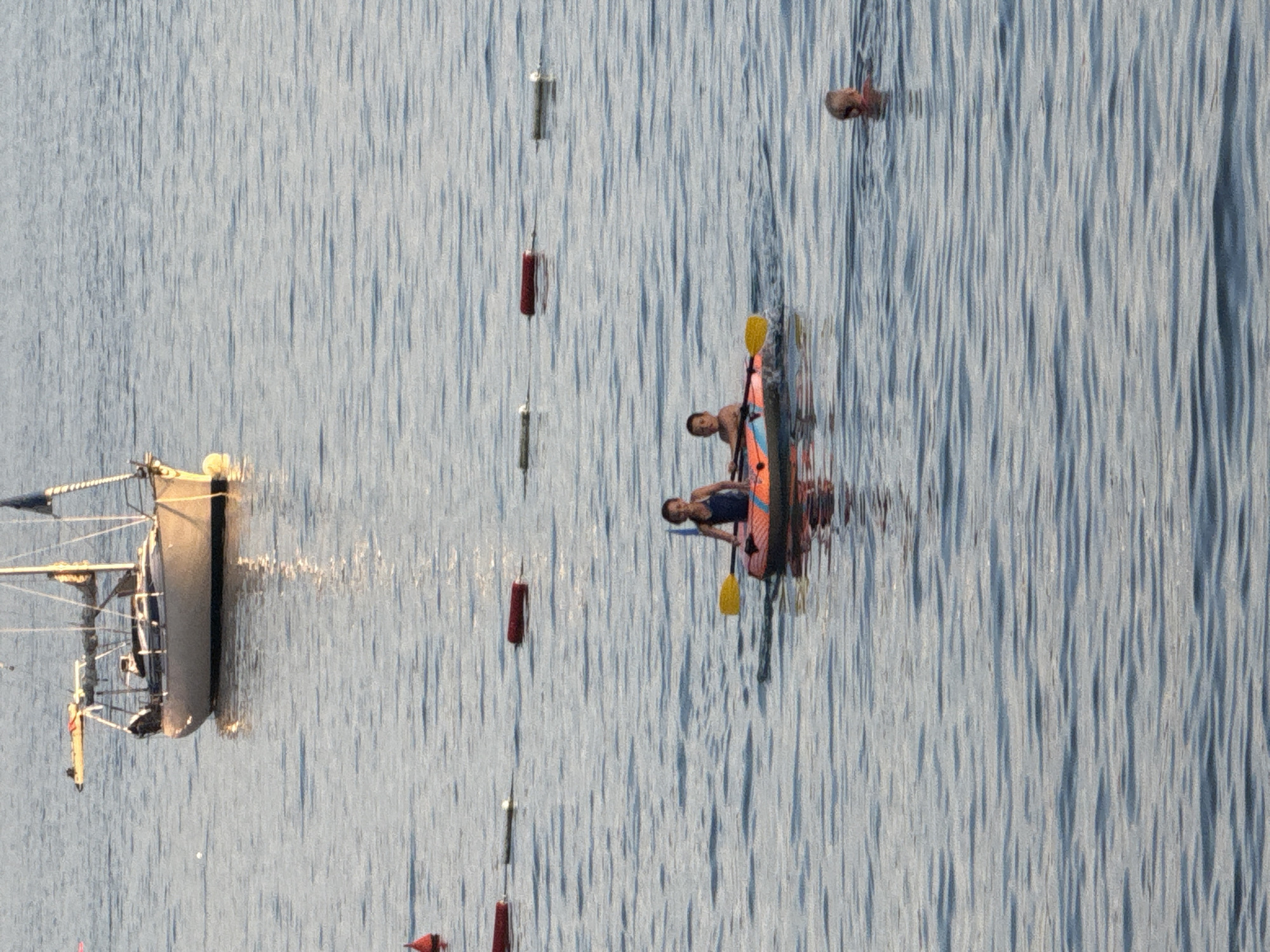 The image, taken from a high vantage point, captures a serene body of water with ripples creating a textured surface. The scene is set outdoors, likely in a coastal or lake area, given the presence of a small sailboat and buoys.
The primary subjects are two individuals in a kayak. One person is paddling, while the other is seated. They appear to be engaged in a leisurely kayaking activity. There are also a few buoys or markers floating on the water, suggesting a designated area or perhaps a racecourse. A partial view of a sailboat's hull and mast is visible on the left side of the image. The reflection of the boat and the kayak is visible on the water's surface, indicating a calm day with minimal wind.
The lighting suggests it is either early morning or late afternoon, with the sun casting a soft glow on the water. The water itself is a dominant element, reflecting the sky and the objects on it. The overall mood is peaceful and tranquil, with the gentle ripples on the water contributing to this feeling.
There is no visible text in the image. The location context of Rafailovići, Montenegro, aligns with a coastal setting where such activities and watercraft would be common.
