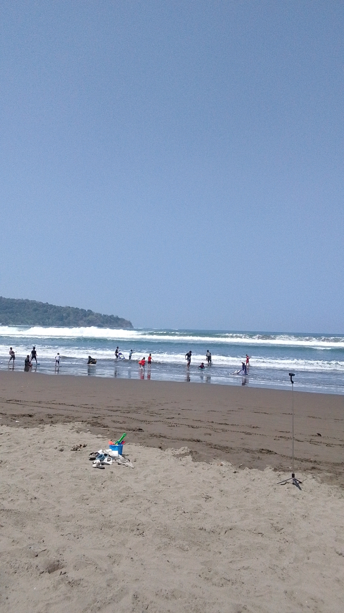 The image depicts a sunny day at Desa Pagandaran, Indonesia, with a beach scene. In the foreground, a pile of beach toys, including a blue bucket with a green shovel and rake, along with a pair of flip-flops, rests on the sand. Further back, several individuals are visible near the water's edge. Some appear to be children playing in the shallow water, while others, possibly teenagers or adults, are wading or holding surfboards. The ocean exhibits moderate waves breaking near the shore, and a distant, tree-covered hillside forms the backdrop. The sky is clear and blue, with no clouds visible. A thin, dark pole with a tripod base stands on the right side of the frame in the sand.