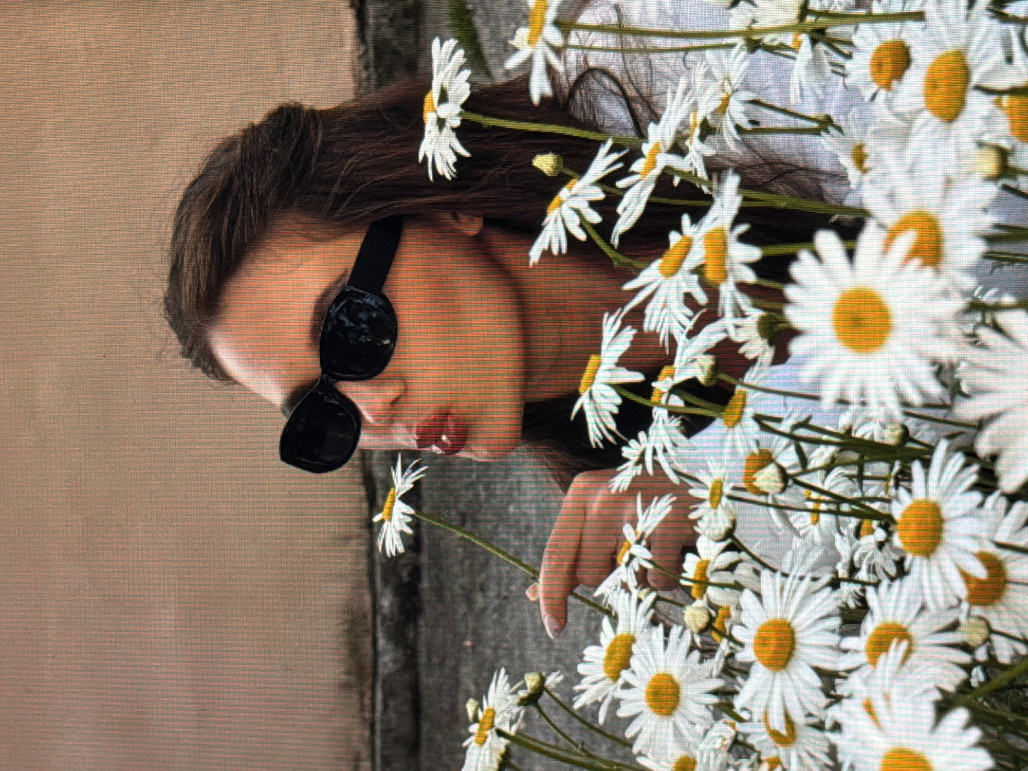 In the city of Sirius, Russia, a woman with dark hair, wearing black sunglasses and a white top, is captured in a close-up shot. She is surrounded by a field of daisies and is holding one of the flowers, bringing it to her lips as if to kiss it. Her lips are painted red, and she is looking slightly to her left. The background consists of a muted, textured wall, suggesting an outdoor urban setting. The lighting is soft, highlighting the details of the woman's face and the surrounding flowers.
