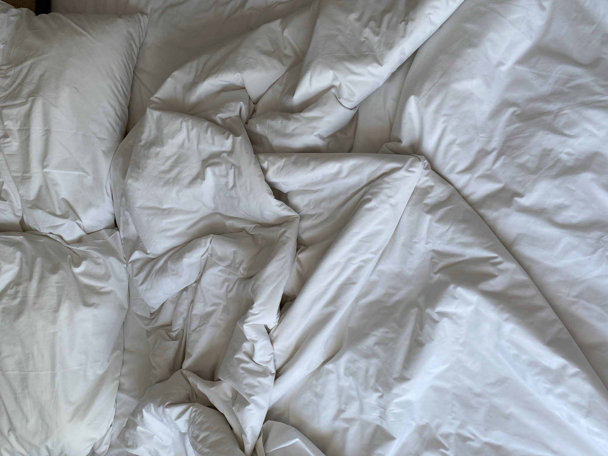 The image shows a rumpled white duvet and two pillows on a bed. The lighting suggests it is daytime, with soft shadows indicating a window is likely nearby. The scene is a close-up, focused on the texture and folds of the bedding. There are no people or animals visible. The setting appears to be a bedroom in a residential or hotel environment, consistent with a location in Nha Trang Ward, Vietnam.
