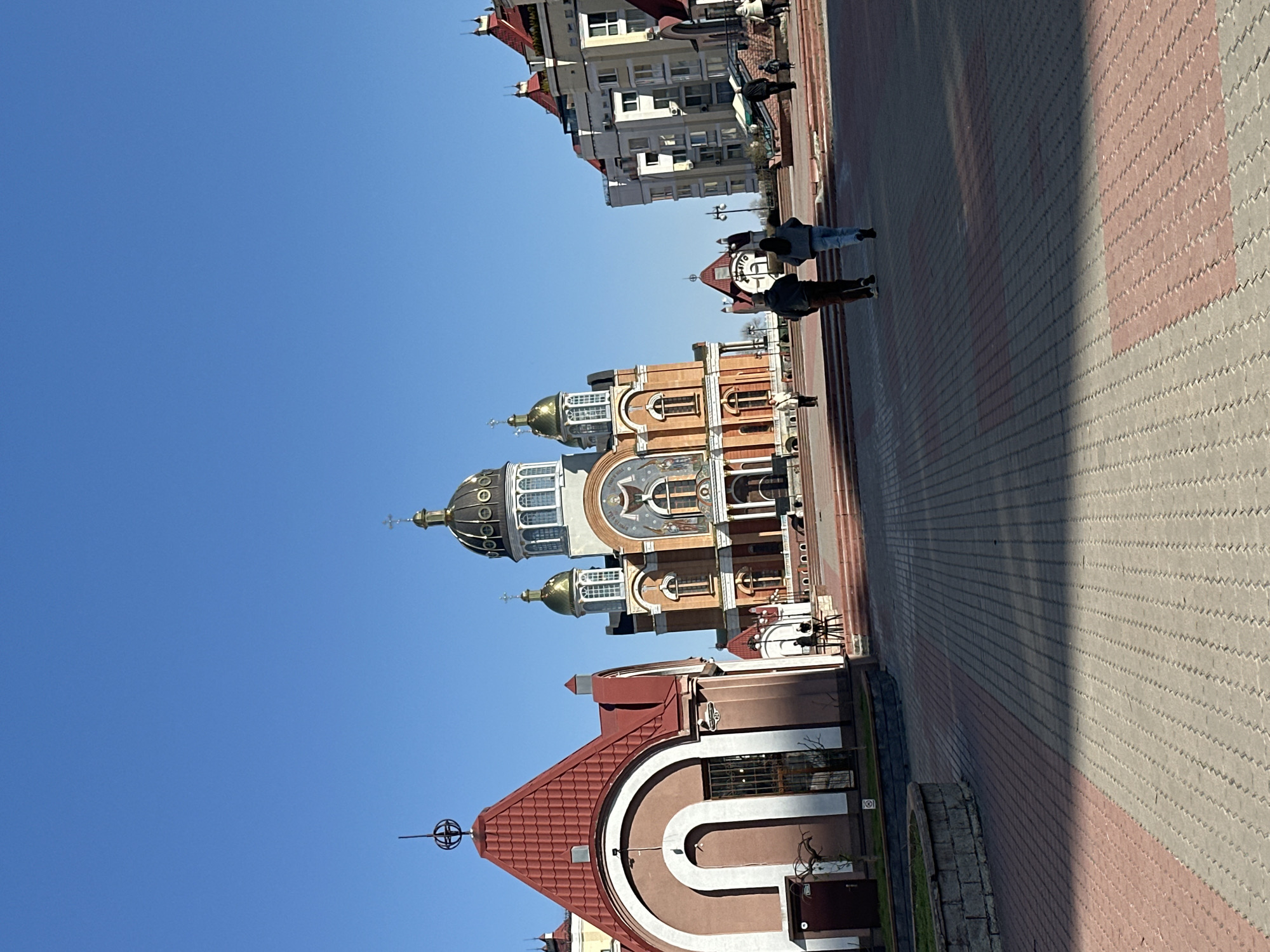 The image shows a view of the city of Kyiv, Ukraine. A large, ornate church with multiple domes stands prominently in the foreground. A paved walkway occupies the right side of the image, with two people walking along it. Buildings line the top of the frame under a clear blue sky.