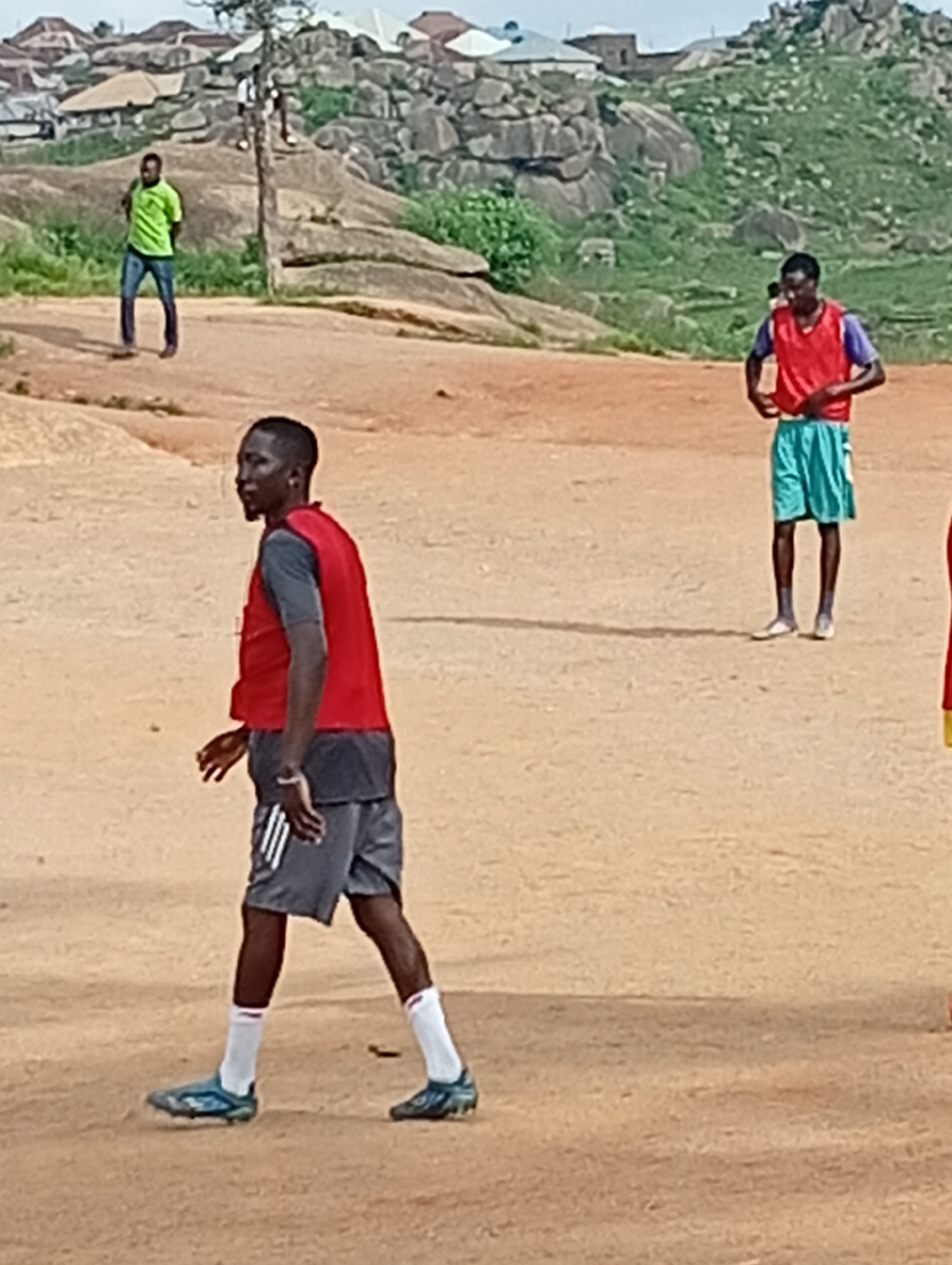 The image shows a group of young men on a dirt field, likely a football pitch, in Jos, Nigeria. In the foreground, a man wearing a red vest over a grey t-shirt and grey shorts with white stripes is looking to his left. He is wearing white socks and blue football cleats. Behind him, a man in a red vest and blue shorts stands with his hands on his hips, and another man in a green t-shirt and blue jeans stands further back. In the background, there are rocky hills with scattered buildings and vegetation, typical of the landscape in Jos. The sky is overcast. The overall impression is of an informal football practice or casual gathering on a sunny day.