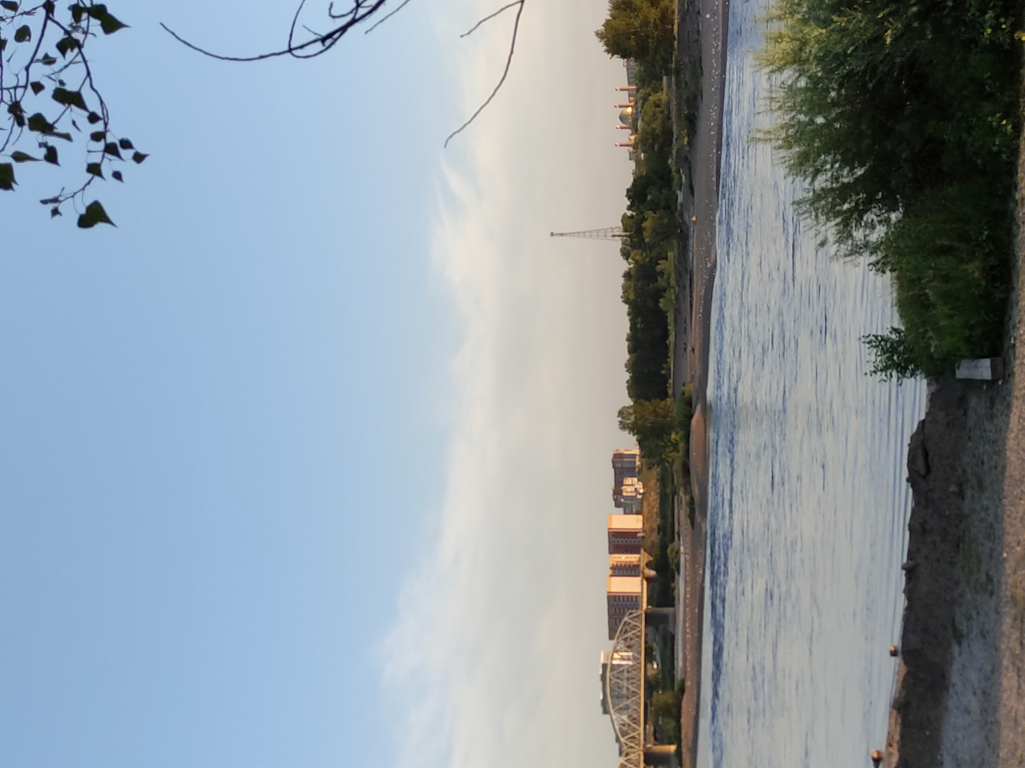 The image shows a wide, calm river flowing through a city. On the far bank, a modern bridge with a metal truss structure spans the water, leading to a cluster of tall, rectangular buildings with colorful facades, appearing to be apartment blocks. Further in the distance, a tall, lattice radio tower stands next to a domed building complex, possibly religious or governmental, with several smaller spires. The foreground features a sandy embankment with scattered rocks and a patch of dense green vegetation. The sky is a clear, pale blue with wispy clouds, and the overall lighting suggests late afternoon or early evening. The scene captures a view across the Irtysh River in the city of Semey, Kazakhstan, showcasing a blend of urban development and natural landscape. There are no people visible in the image.
