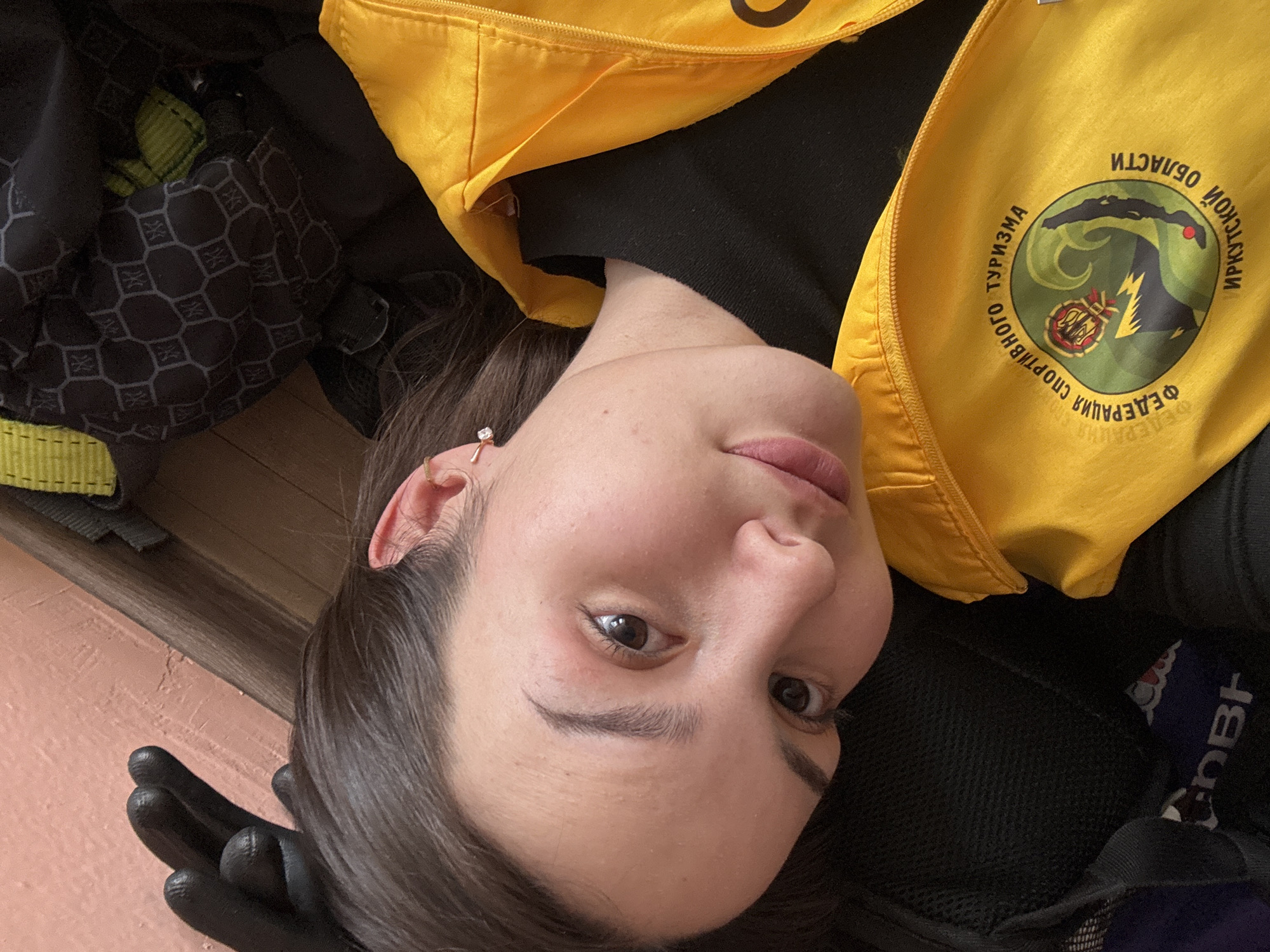 A female individual is depicted in a close-up, angled shot, appearing to recline indoors. She has dark hair, a fair complexion, and brown eyes, looking directly at the camera with a slight expression. Her right ear displays a small, light-colored stud earring. A black glove covers her partially visible left hand, positioned near her head. She wears a black long-sleeved garment underneath a bright yellow zippered vest. The vest features a circular emblem on the left chest, displaying the Russian text "ФЕДЕРАЦИЯ СПОРТИВНОГО ТУРИЗМА ИРКУТСКОЙ ОБЛАСТИ" (Federation of Sports Tourism Irkutsk Region), encircling a multi-colored logo depicting a mountainous landscape. Partially visible behind her are a black object with a mesh texture, and another dark object with a geometric hexagonal pattern. A light orange wall and a segment of a wooden floor or trim are visible in the background. The scene is consistent with an indoor setting within the Irkutsk Region, Russia, given the vest's insignia; however, the precise location within the city of Tayshet, Russia, is not discernible from the image.