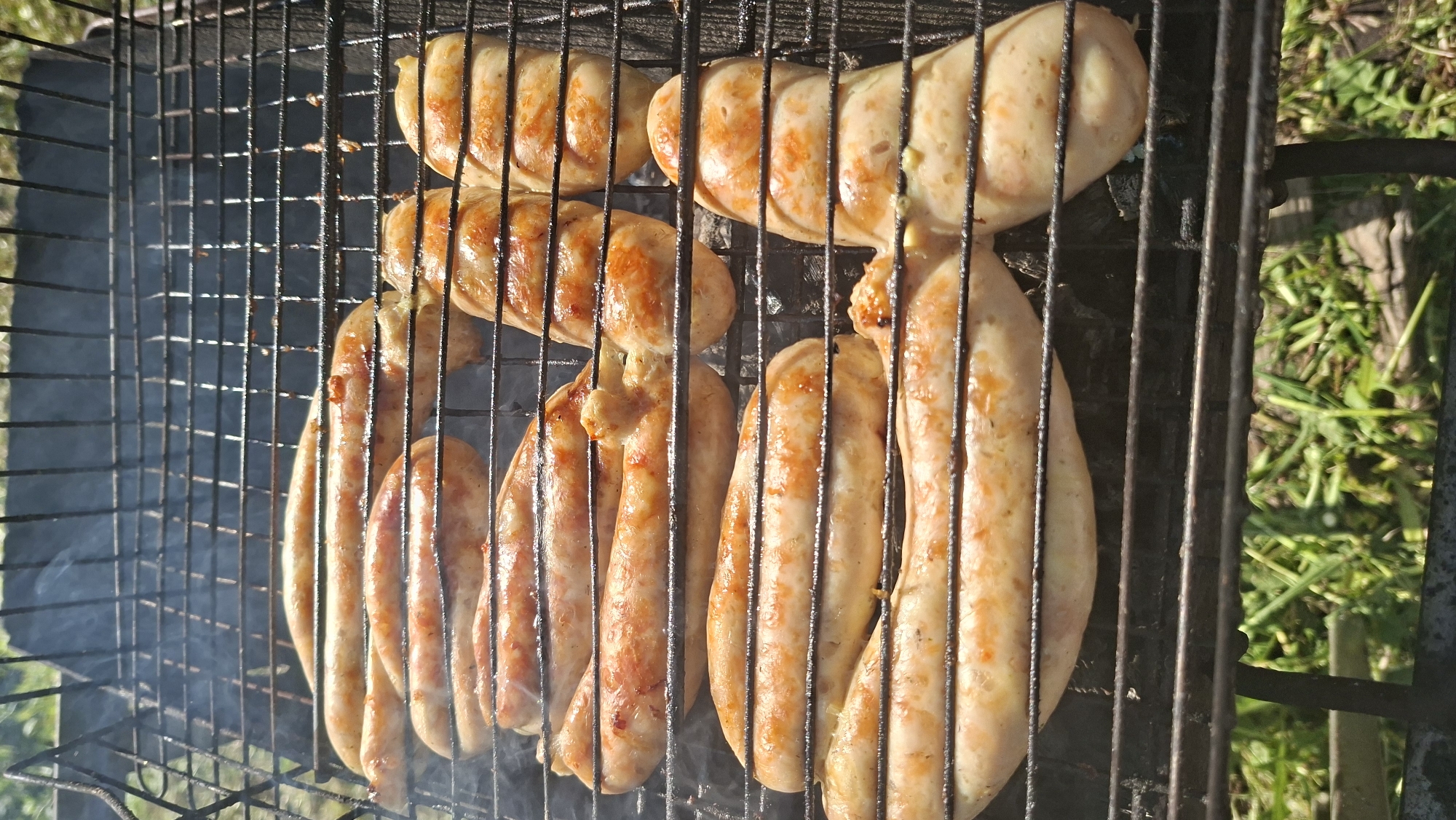 The image shows several sausages grilling on a metal grill. Smoke is rising from the grill. The sausages appear to be partially cooked with some browning visible. Some grass is visible around the grill. The location is near Сваткаўскі сельскі Савет, Belarus.