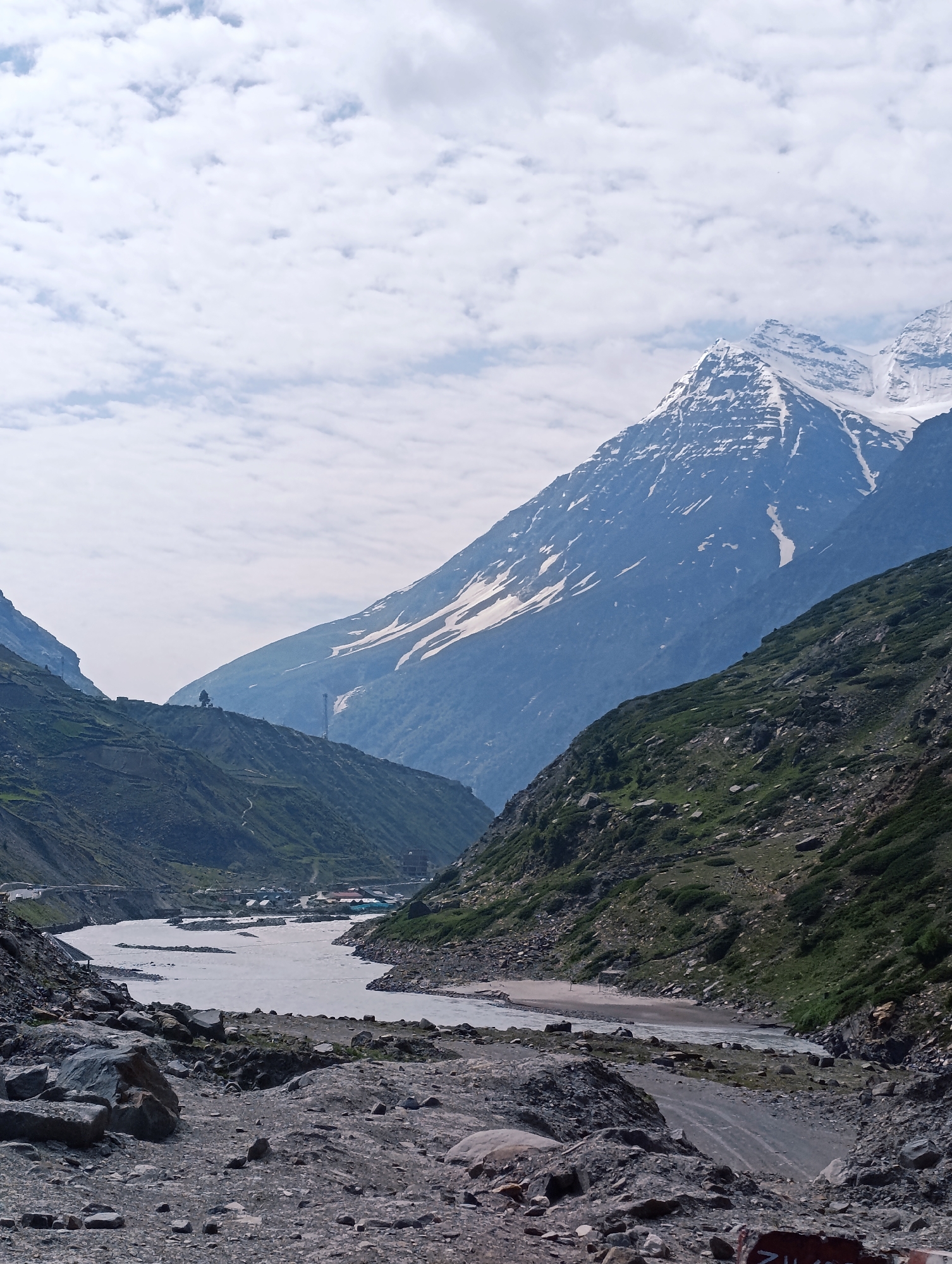 The image depicts a scenic view of the Lahaul Valley in Sissu, India, featuring a meandering river flowing through a valley flanked by steep, rocky mountains. The mountains on the right are covered in patches of snow, with their peaks disappearing into a cloudy sky. The slopes of the mountains on the left are greener, with some evidence of terraced farming and scattered structures. The foreground is dominated by a rocky, uneven terrain, with a dirt road visible on the right side. The river is wide and appears to be carrying a significant amount of water. The overall impression is one of natural beauty and the ruggedness of the Himalayan landscape.