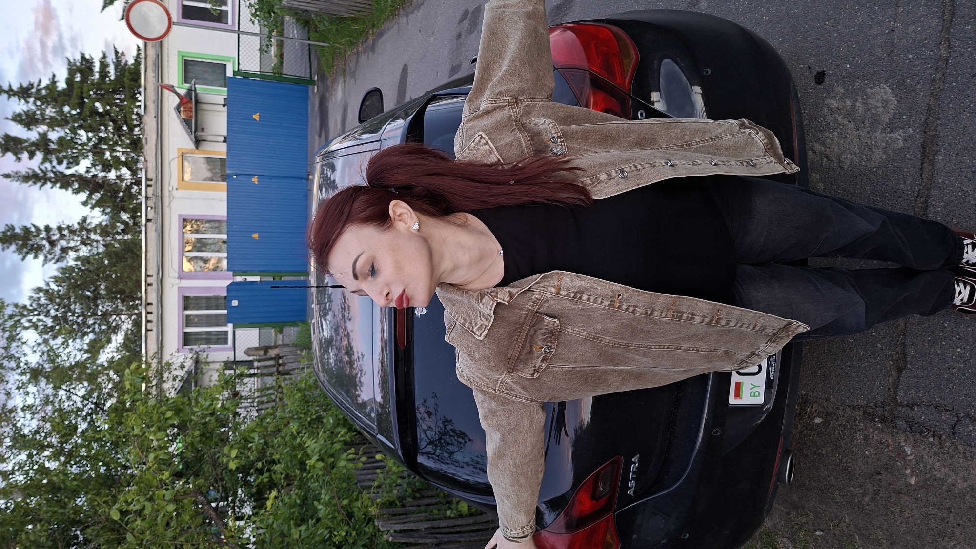 A woman with long, red hair styled in a ponytail is lying on the back of a black Opel Astra car. She is wearing a black t-shirt, dark jeans, a tan denim jacket, and red and black sneakers. The car has a Belarusian license plate. The backdrop includes a building with a blue fence and multicolored window frames, trees, and a street. The location is Забалоцкі сельскі Савет, Belarus.
