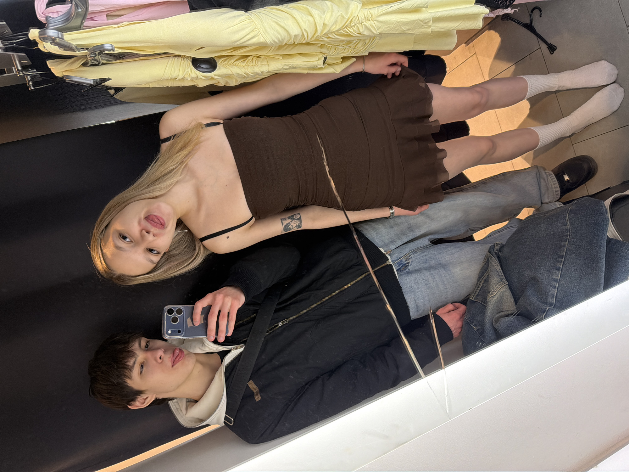 A young couple is taking a selfie in a likely indoor location. The woman has blonde hair, a brown dress, and white socks. The man has dark hair, is wearing a black jacket with a white hooded undershirt, blue jeans, and is holding a phone. The setting appears to be in or near a clothing store, with clothes hanging on a rack in the background. The location is Yekaterinburg, Russia.