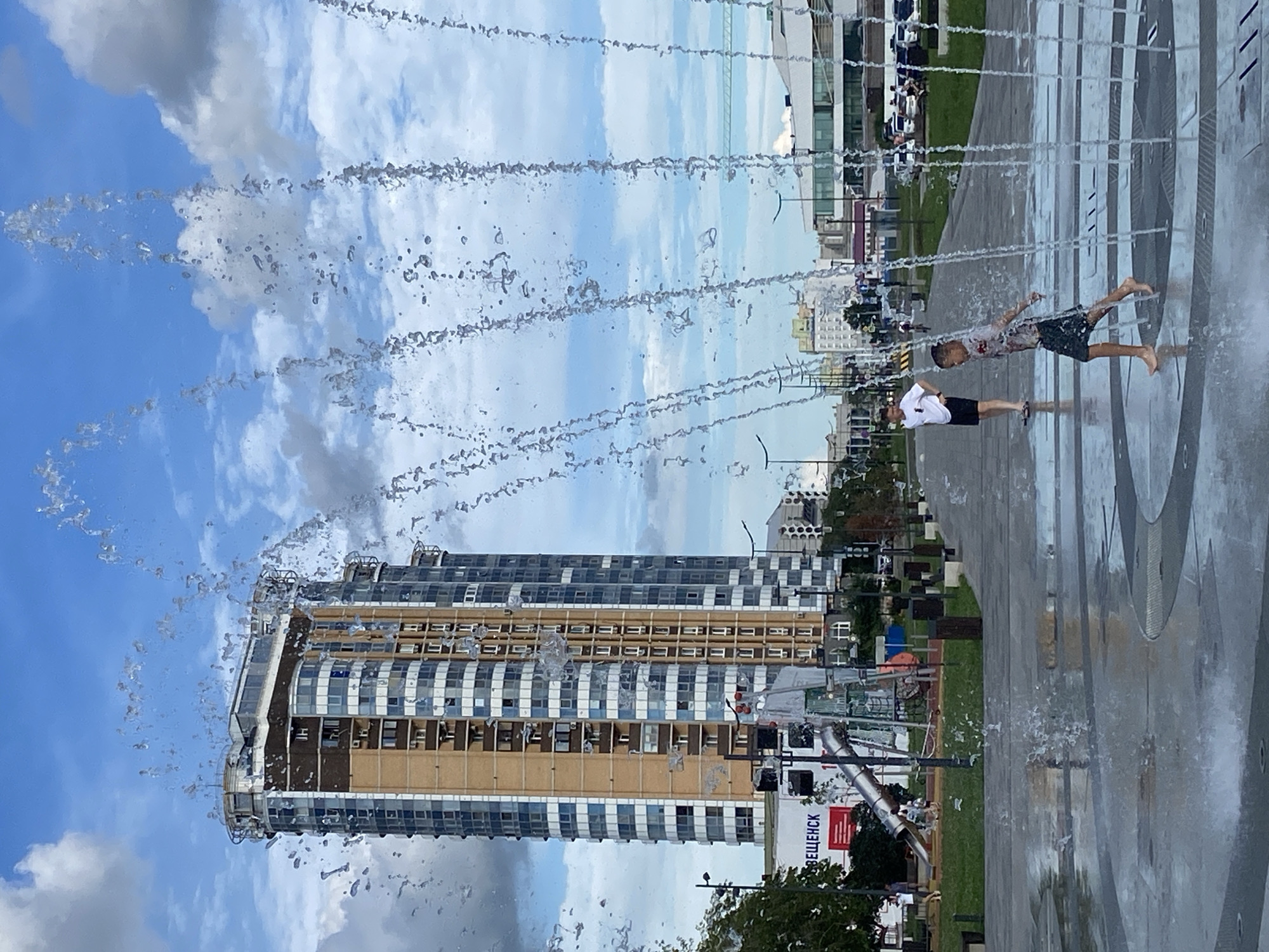 In Blagoveshchensk, Russia, on a cloudy day, a boy runs barefoot through a water fountain in a public plaza. The plaza features a modern, tall, multi-story building with a rounded facade and numerous windows, and a playground with a metal slide and climbing structures. Lampposts are visible along the paved walkway, and cars are parked in the background. The boy, wearing a patterned t-shirt and dark shorts, is captured mid-stride, surrounded by the spray of the fountain. Another young male, wearing a white t-shirt and black shorts, stands nearby observing. The overall scene depicts a recreational area in an urban environment.