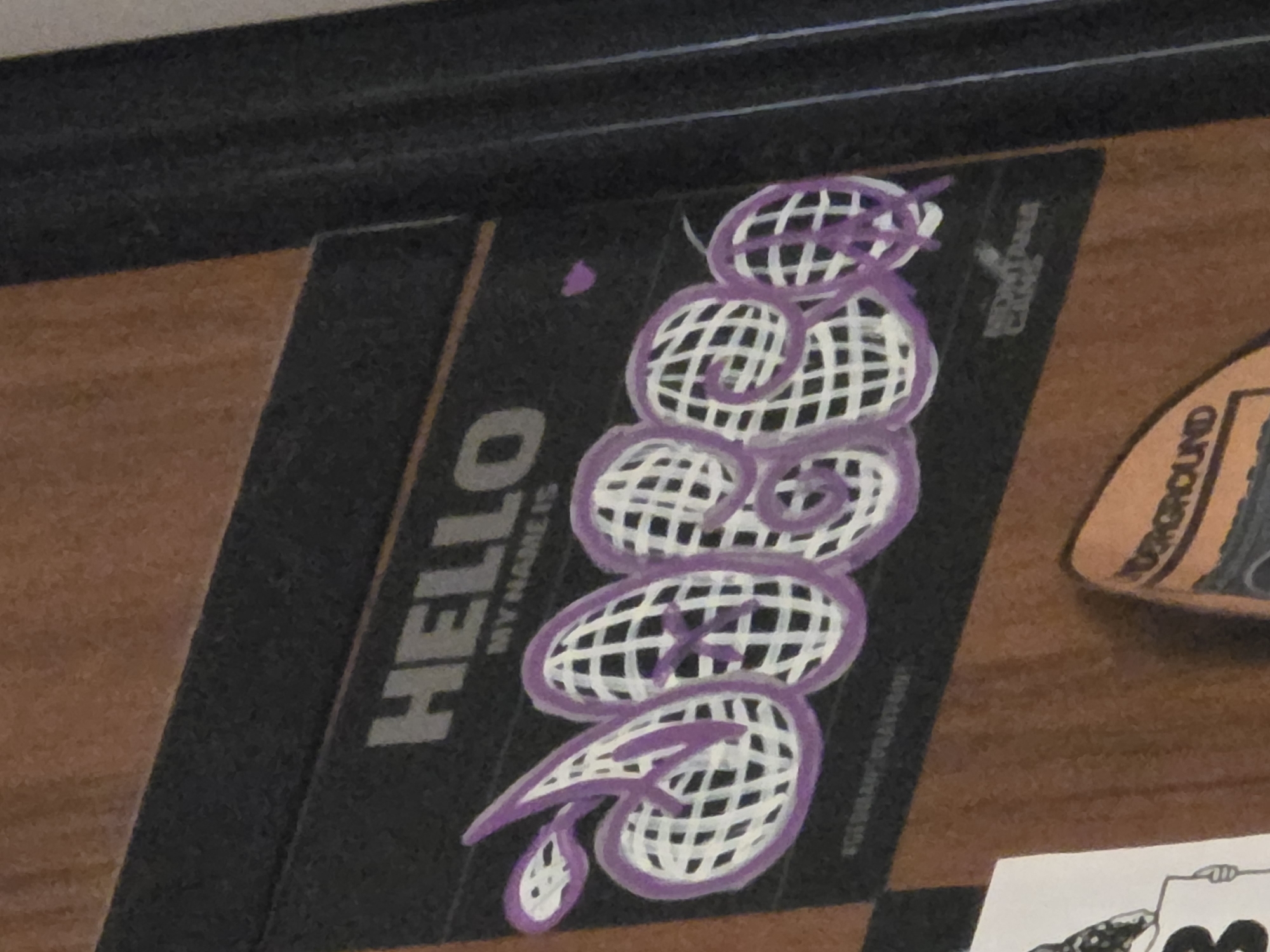 The image shows a close-up of a black sticker with graffiti art on a wooden surface. The sticker has "HELLO" written in large, bold, grey letters at the top. Below that, in smaller grey letters, it says "MY NAME IS". The main feature of the sticker is a graffiti tag in purple and white, which appears to spell out "DOBO" or a similar name. The letters are stylized with a grid pattern inside and outlined in purple. There are small purple shapes scattered around the graffiti. In the bottom right corner of the sticker, smaller text reads "MONTANA CANS". Another small sticker with the word "GROUND" is visible at the bottom. The background appears to be a wooden door or paneling.