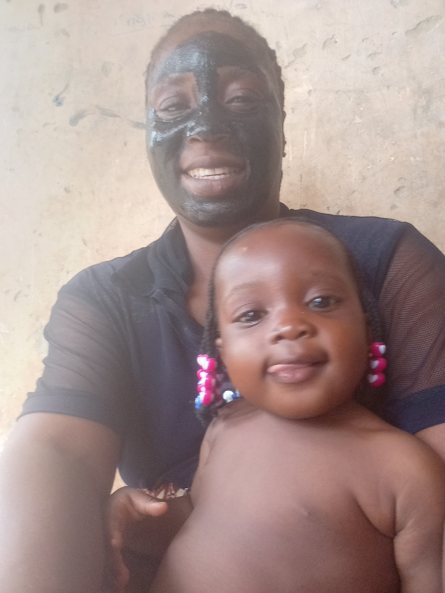 A selfie shows a woman with a black facial mask smiling while holding a baby who is sticking out its tongue. The baby has braids with pink and white beads on the ends. They appear to be indoors in front of a tan wall. The location in Akwanga, Nigeria, is suggested but unconfirmed by visual evidence.