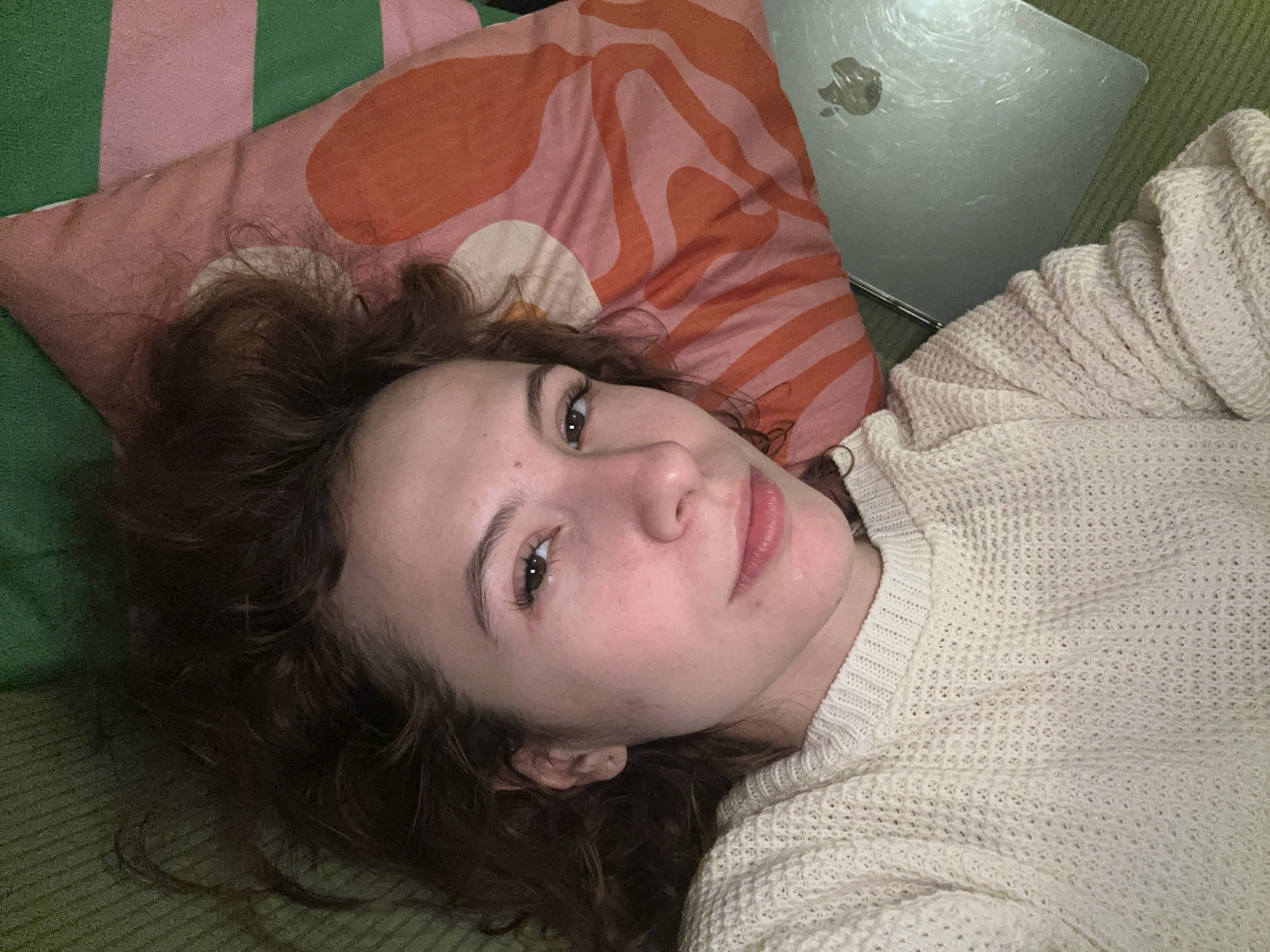 A young woman is lying down indoors, looking directly at the camera with a calm and gentle expression, hinting at a slight smile. Her head rests on a decorative pillow featuring an abstract pattern of swirling orange and white shapes on a pink surface, set against a larger background of green and pink vertical stripes.
She has dark brown, curly hair that frames her face and flows onto the pillow. Her eyes are light brown or hazel, and her long, dark eyelashes are prominent. She is wearing a light-colored, possibly off-white or cream, knitted sweater with a textured, open-weave pattern.
To the right side of the frame, behind her head and shoulder, a silver laptop with a visible Apple logo is partially seen, reflecting light. The surface she is lying on appears to be a soft green fabric, possibly a couch or bed. The lighting in the image is soft and ambient, suggesting an indoor setting, possibly in the evening or under subdued light conditions. The scene conveys a sense of relaxation and comfort. The location context provided is Novi Sad, Serbia.