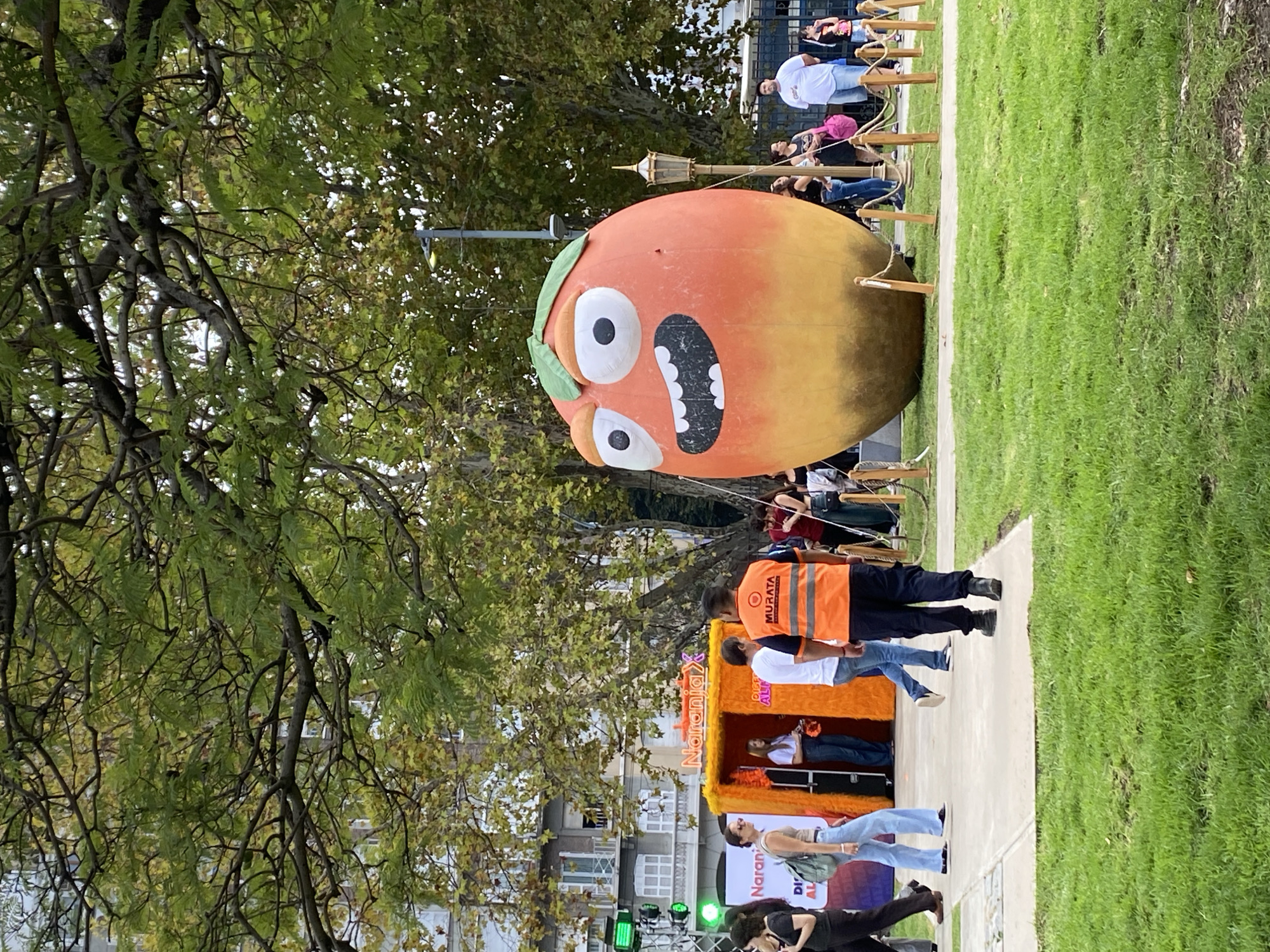 The image shows a public event in Buenos Aires, Argentina. A large, inflatable, orange-shaped cartoon figure with a surprised facial expression dominates the scene. The figure stands on a street near a green lawn. A crowd of people stands behind a low wooden fence in the background. In the foreground, a temporary stage with orange decorations is visible, and people are standing around it. One person wears an orange vest with the word "MURATA" on it. The street is lined with buildings, and a tree with green foliage is visible on the left side of the image. The lighting appears to be from daylight. Traffic signals are visible in the background.