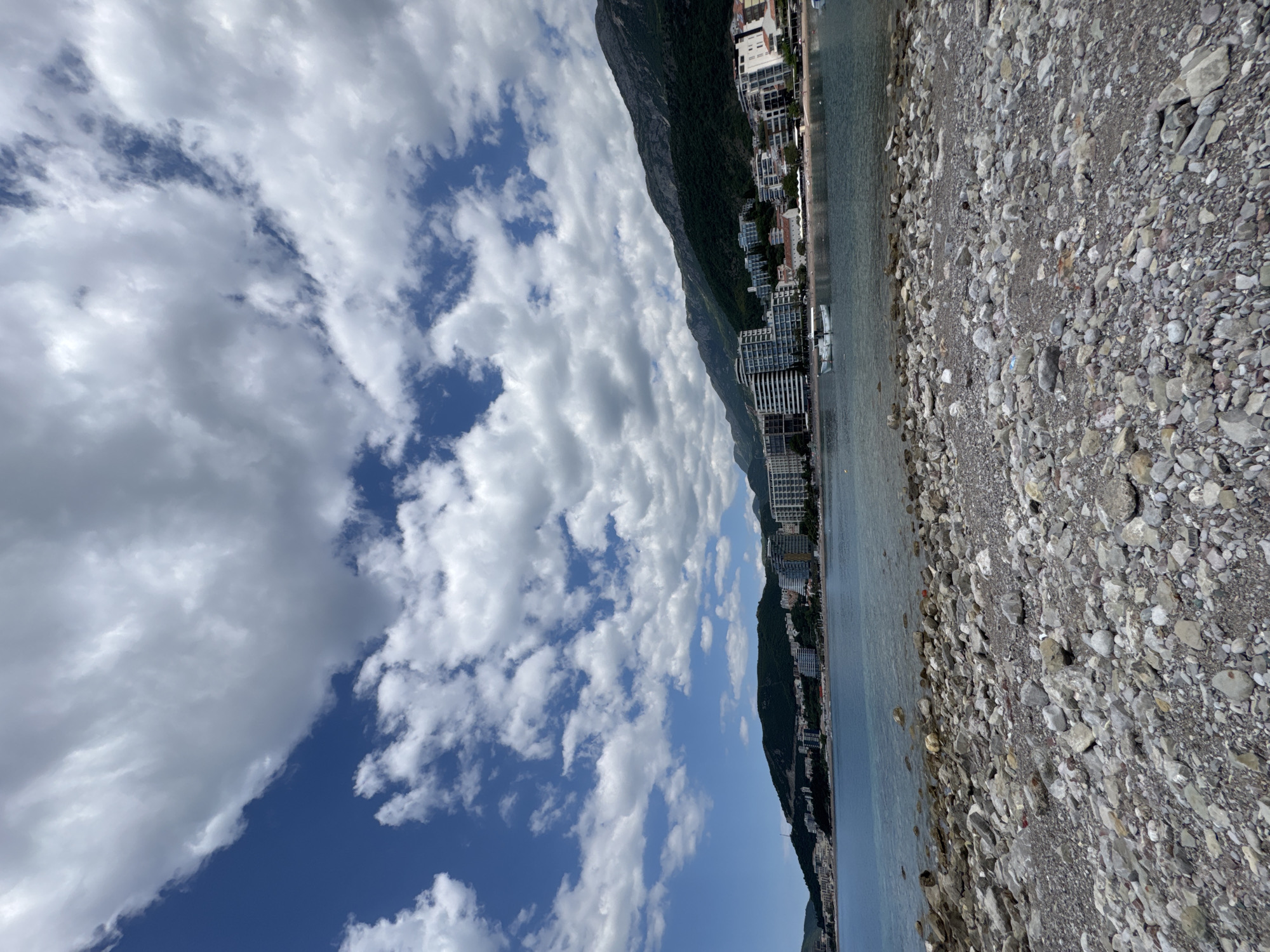 The image captures a view of Rafailovići, Montenegro, from the beach, oriented vertically. The right portion shows a pebble beach with stones of various sizes and shades of gray. Adjacent to the beach, the Adriatic Sea extends toward the center. Beyond the water, the urban landscape of Rafailovići is visible with a line of buildings along the coast, appearing modern and high-rise in parts. Behind the buildings rises a steep, green, tree covered mountain ridge. The sky dominates the left portion of the image with fluffy white clouds against a blue backdrop. The overall impression is of a summer day at the coast, showcasing the natural and built environment of the coastal town.
