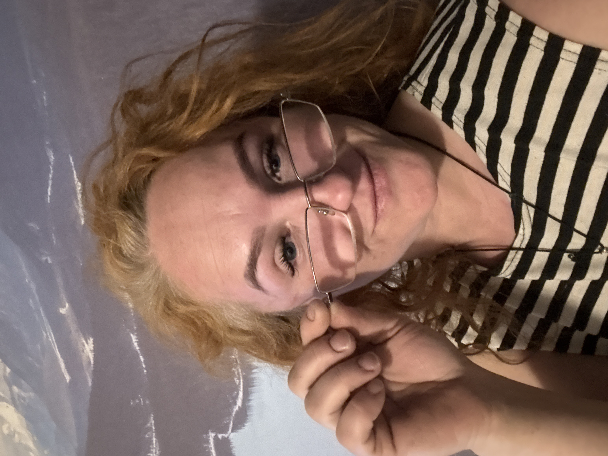 A fair-skinned woman with wavy red hair is holding her glasses on her face with her right hand. She is wearing a black and white striped shirt. The background is a mottled gray-blue surface. There is no indication that the image was taken in Chaikovsky, Russia.