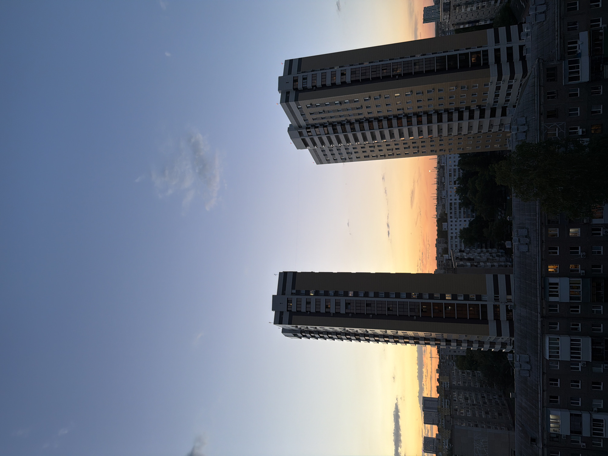 The image shows two tall buildings in an urban environment at dusk. The sky is a gradient of blue to orange. The buildings are multi-story with numerous windows, some of which are illuminated. Other buildings are visible in the background.