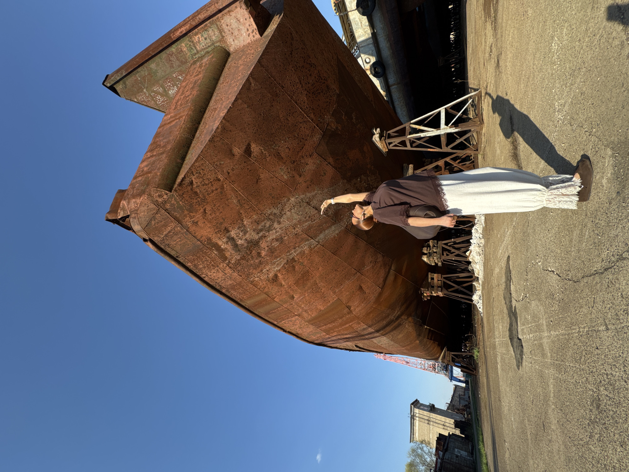 The image captures a scene in what appears to be a shipyard or industrial area. A large, rusted metal hull, likely of a ship, dominates the left side of the frame. The hull is positioned in such a way that it appears to loom over the viewer. A woman stands next to the ship, seemingly touching the metal. She is wearing a dark brown top and a long, white skirt or dress with lace details at the bottom. She is also wearing brown shoes. Her right arm is extended toward the metal hull.
The background includes a clear blue sky, contrasting sharply with the rusted metal. To the right, the ground is paved. There are also wooden structures, possibly supports for the ship. In the far background, some buildings and a crane are visible.
The city of Chaikovsky, Russia, is not explicitly confirmed within the image but is provided as part of the context. It is possible that this shipyard or industrial area is located within or near Chaikovsky.