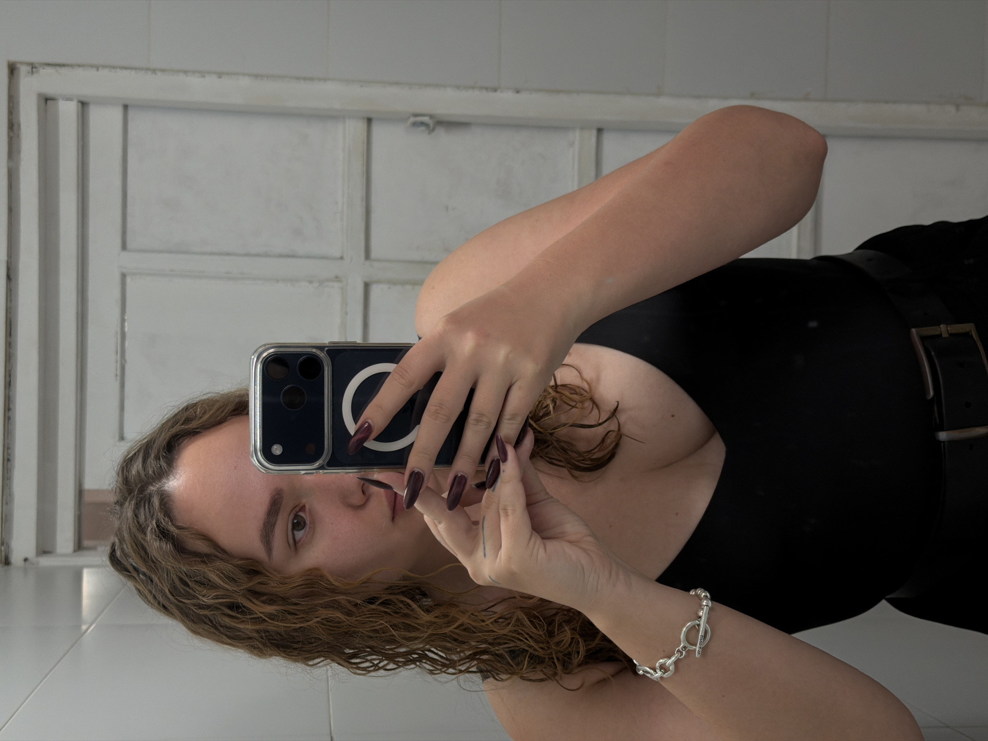 A woman is taking a selfie in a bathroom. She is holding a phone with a dark case and a circular magnet on the back, covering part of her face. She has curly hair, light skin, and dark nail polish. She is wearing a black top and a belt with a large buckle. A silver bracelet is visible on her right wrist. The bathroom has white tiled walls and a white-framed window or door in the background. There is nothing in the image to indicate this is in Ramat Gan.
