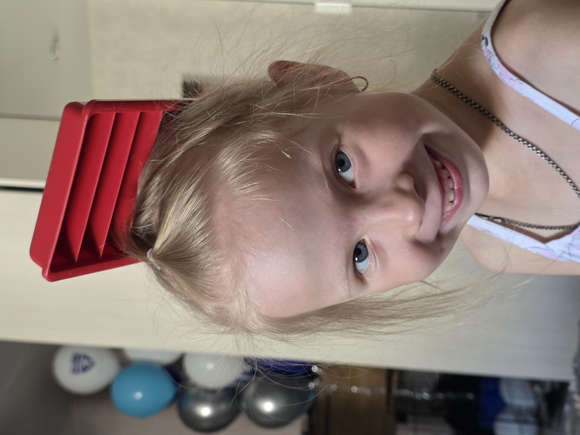 A young girl with blonde hair and blue eyes is pictured with a red plastic object stuck in her hair. She is smiling at the camera. In the background, there are several balloons of various colors, including white, blue, and silver. The setting appears to be indoors, with a light-colored wall and some furniture visible. The image does not provide any specific location information within the city of Chaikovsky, Russia.