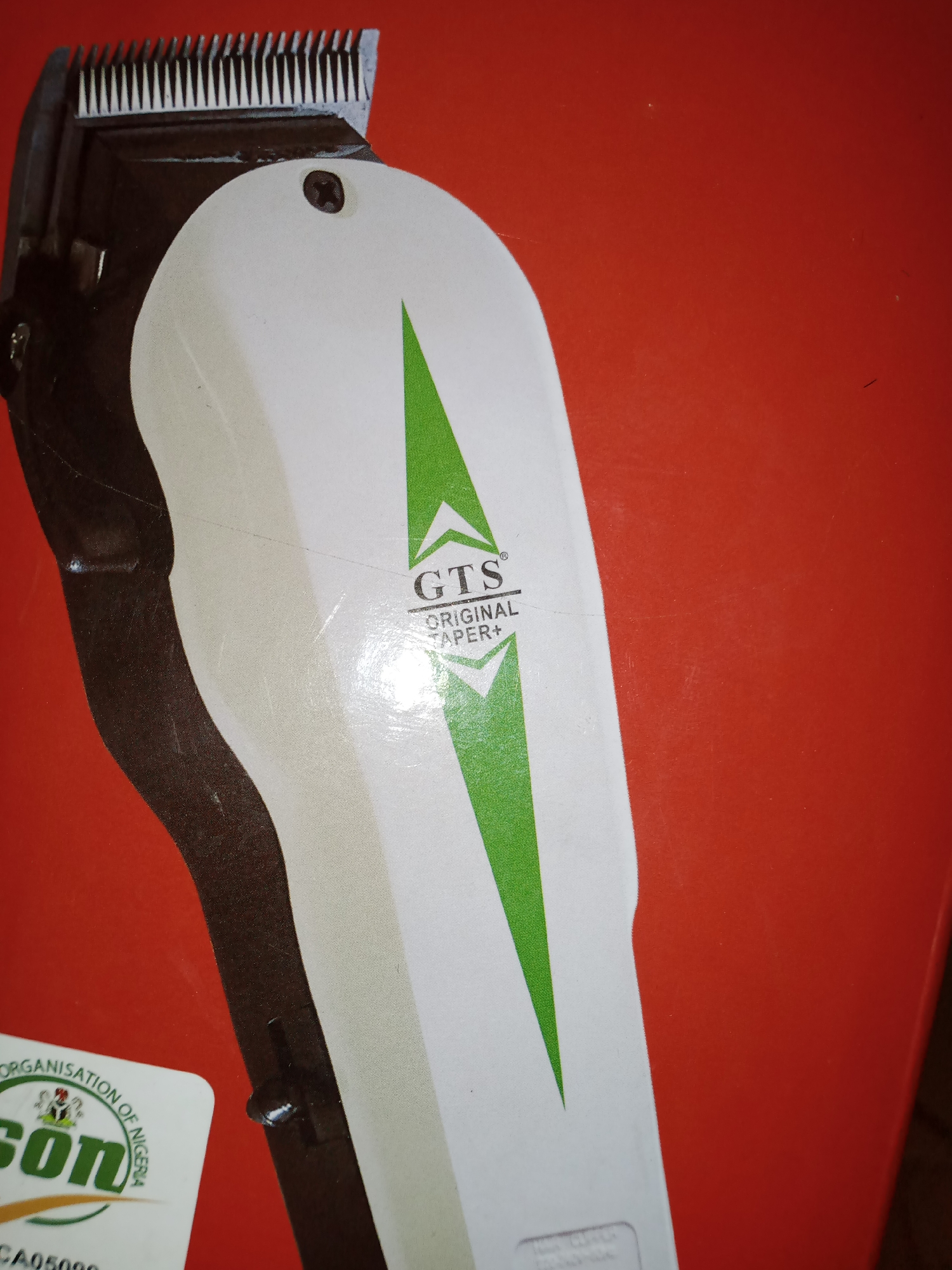 The image shows a close-up of a white and black hair clipper with a green triangular logo. The logo has the text "GTS ORIGINAL TAPER+" on it. Below the clipper, a portion of a sticker is visible with the text "ORGANISATION OF NIGERIA" and "son". The background is a solid red color. The scene does not provide any information about the city of Jalingo, Nigeria, or any people, actions, or interactions.