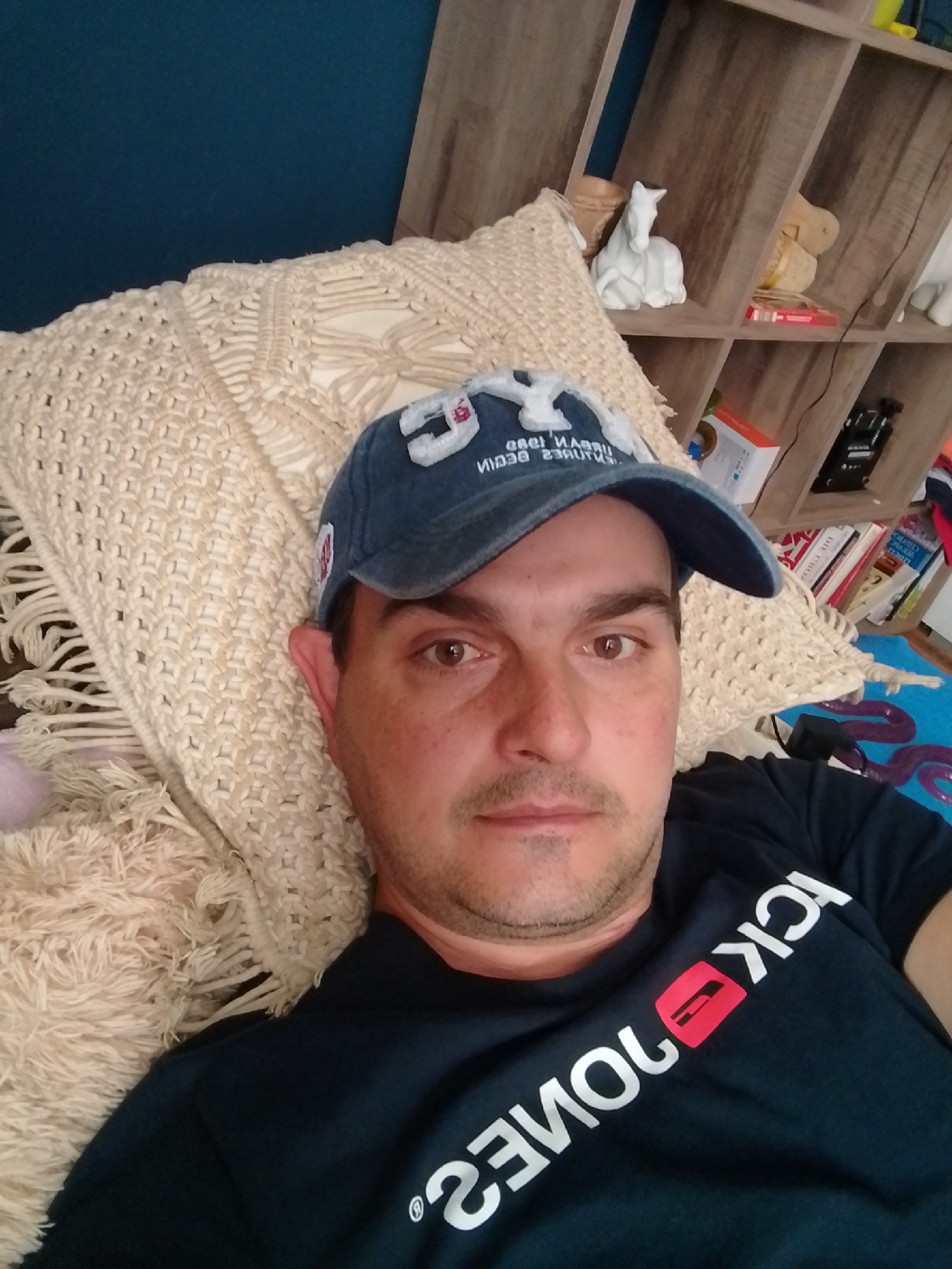 A man wearing a blue baseball cap and a black t-shirt with white lettering that reads "JACK & JONES" is lying down on a beige, macrame-style cushion with fringe. The man is looking at the camera. Behind him, a wooden shelf unit is visible, filled with various items including books, a white horse figurine, and other decorative objects. The overall environment appears to be indoors, possibly a living room or bedroom. There is no specific information in the image that would suggest a location in the city of Wedel, Germany.