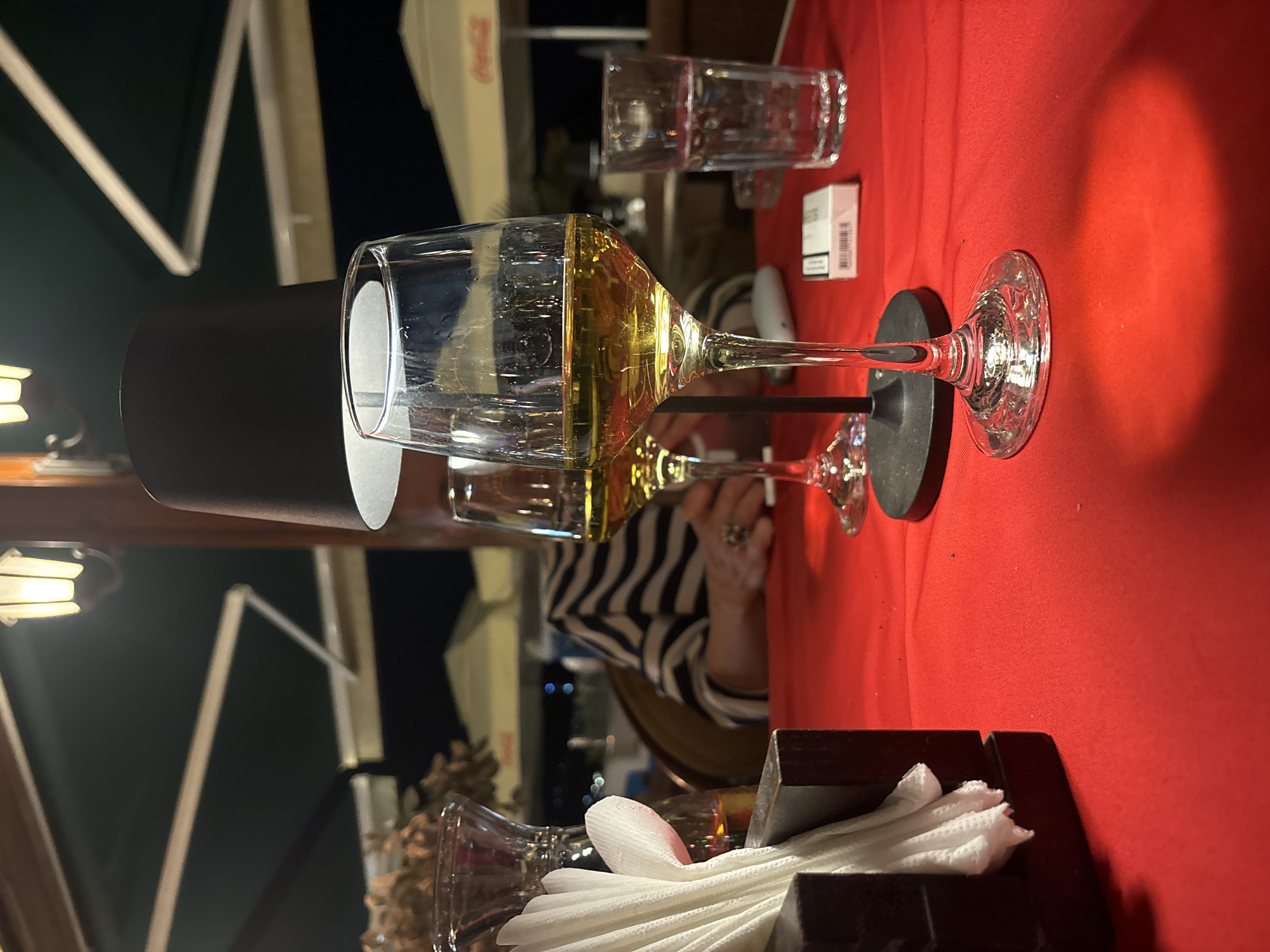 The scene is set at an outdoor restaurant, likely in Budva, Montenegro, given the coastal architecture visible. The table is covered with a bright red tablecloth. Several wine glasses, filled with a golden liquid, possibly wine, are prominently displayed. Some glasses are empty. There is a black table lamp near one of the glasses. Behind the wine glasses, a woman wearing a striped shirt can be seen. There's a packet of cigarettes and water glasses present. Napkins are contained within a dark, wooden container. Elements of outdoor lighting and architectural features suggest a relaxed, evening atmosphere.
