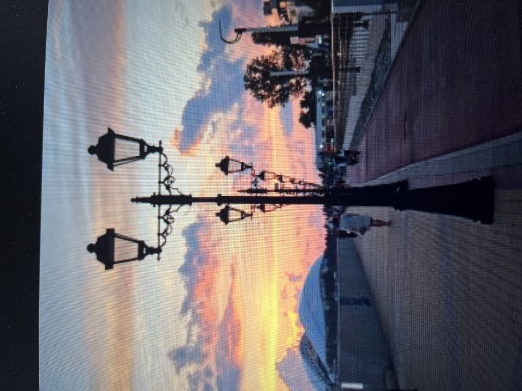 An outdoor, eye-level, full shot shows a street lamp against a sunset sky on a promenade in Khimki, Russia. The sky displays hues of yellow, pink, and orange with scattered clouds. The street lamp is a tall, black, ornate fixture with two lanterns at the top and two additional lanterns extending from the central pole. The promenade is paved with light-colored bricks on the left and a red path on the right. A woman wearing a light-colored dress walks away from the camera on the left side of the promenade. Several indistinct figures are visible further down the promenade, some appearing to be on bicycles or scooters. In the background, there are buildings, trees, and a structure with a white, curved roof, possibly a dome or stadium. The scene captures a moment of dusk along a waterfront or recreational area.