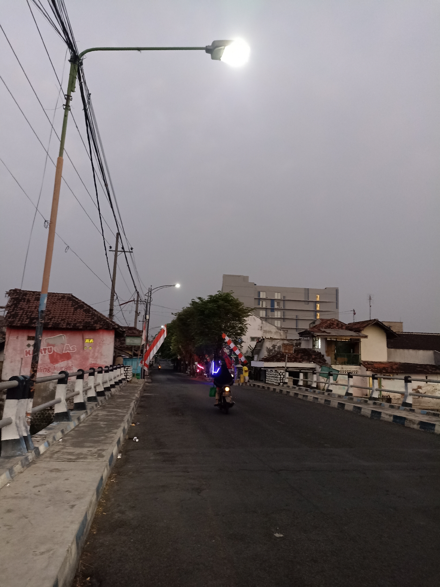 This is a vertical, outdoor, medium-shot photograph taken at dusk on a street in Lumajang, Indonesia. The scene depicts a road with a pedestrian walkway on the left. A street lamp is illuminated overhead, casting a bright light. A motorcycle, ridden by a person whose body is mostly obscured by lights, is moving away from the camera down the road. The road is lined with buildings on both sides, some with traditional tile roofs and others that are more modern. A large, multi-story building is visible in the background. On the left side of the road, a building with red paint and white text, possibly advertising, is partially visible. Several Indonesian flags are displayed along the roadside. The sky is overcast and appears to be a light gray color, suggesting twilight or a cloudy day. The overall atmosphere is that of a street scene during the transition from day to night.