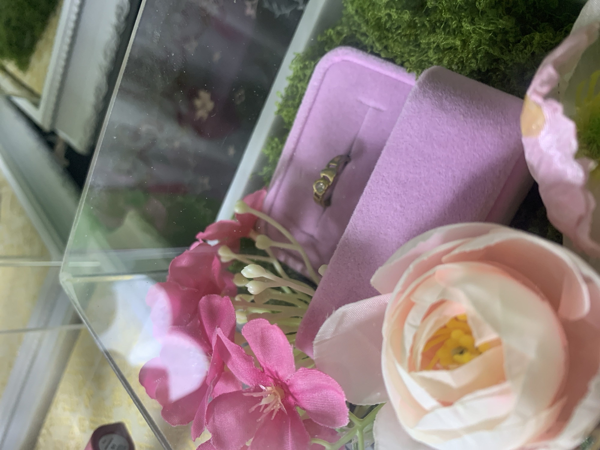The image shows a close-up view of a glass display case containing decorative items. In the foreground, a light pink and white artificial flower is prominently featured. Behind it, to the right, a pink velvet ring box is open, revealing a gold ring with a small clear stone. Further back, pink artificial flowers with small white buds are arranged. The background consists of vibrant green artificial moss. The display case appears to be part of a larger arrangement, with reflections of other objects and a patterned surface visible in the glass. The context suggests this might be a display for wedding or engagement items, possibly in a shop or at an event. There is no discernible indication of the specific location within Kuala Pembuang, Indonesia.