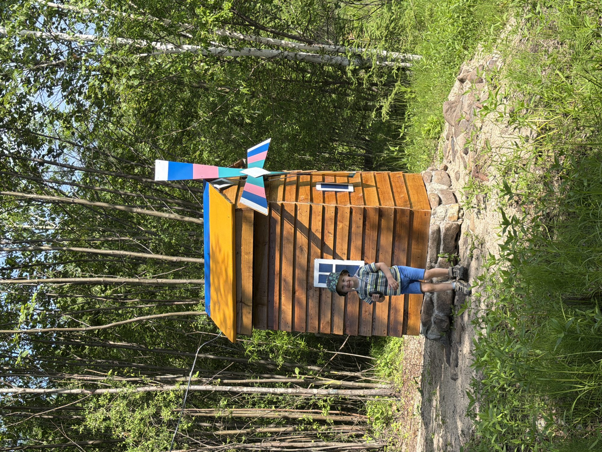 A young boy with a bucket hat, striped t-shirt, and blue shorts stands in front of a small, wooden windmill structure. The windmill has a yellow roof with a blue trim and a colorful pinwheel. The structure is surrounded by rocks and sandy ground, with lush green vegetation and trees in the background. The scene appears to be in a park or outdoor recreational area in Kul'dur, Russia, evidenced by the birch trees and the overall natural setting. The lighting suggests it is daytime, with sunlight filtering through the trees.